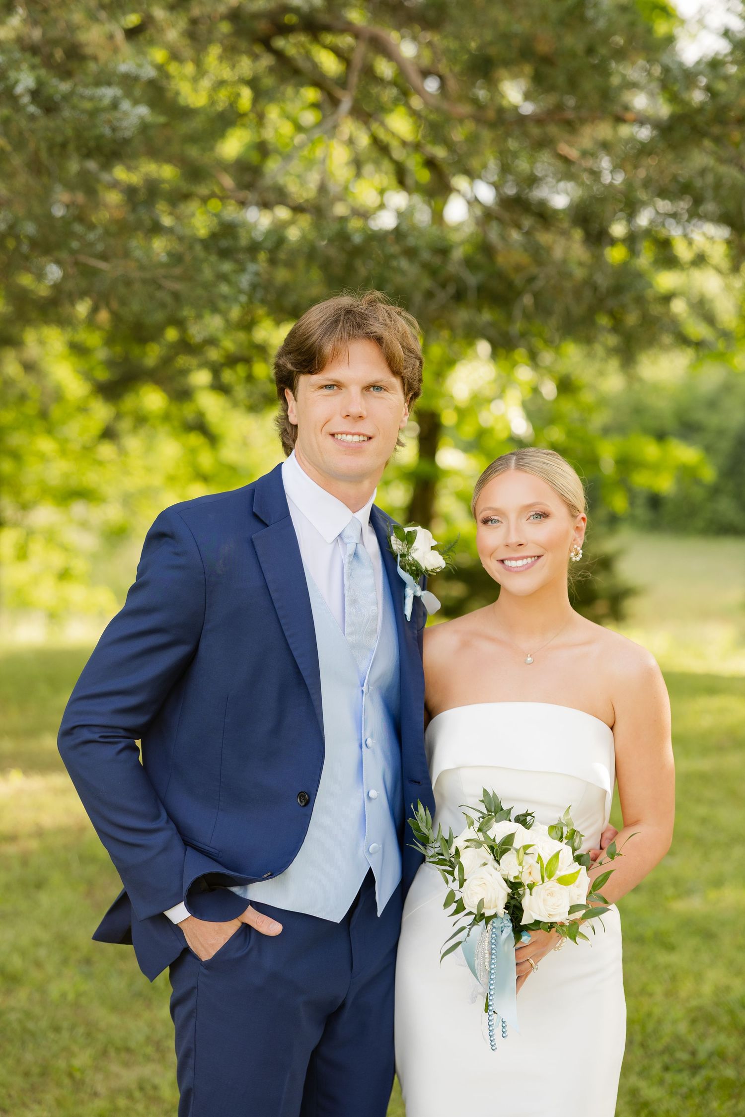 Wedding couple poses outdoors against natural greenery in navy suit and white gown.Cold Springs Events
