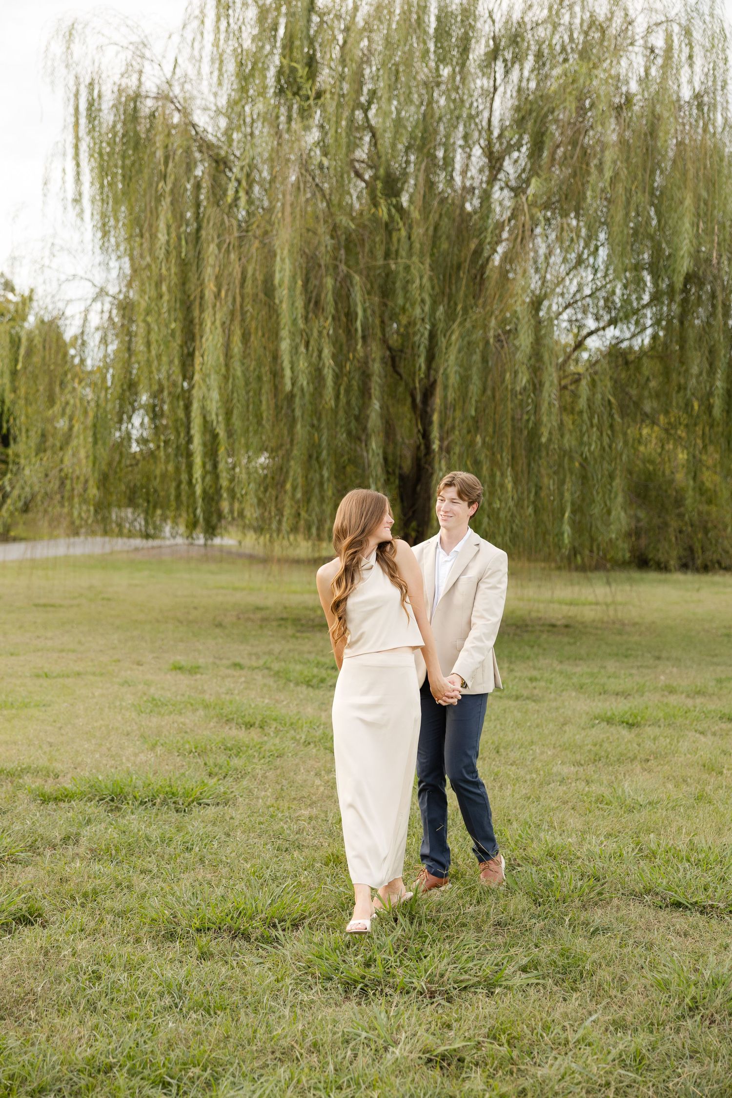 A couple stands together on grass beneath a graceful weeping willow tree in an outdoor setting.