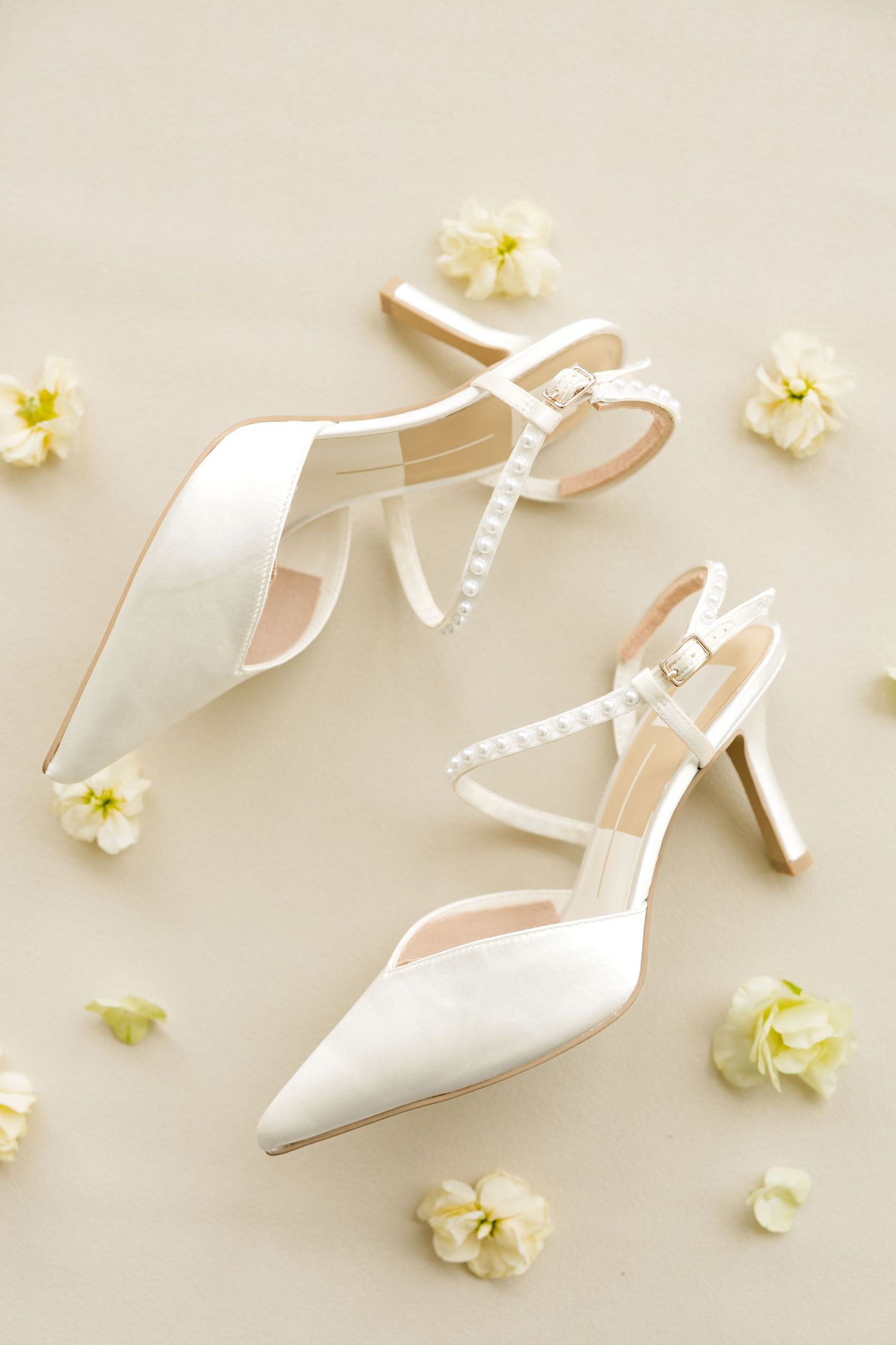 Elegant white satin pointed-toe heels with pearl ankle straps surrounded by delicate cream flowers on a neutral background.