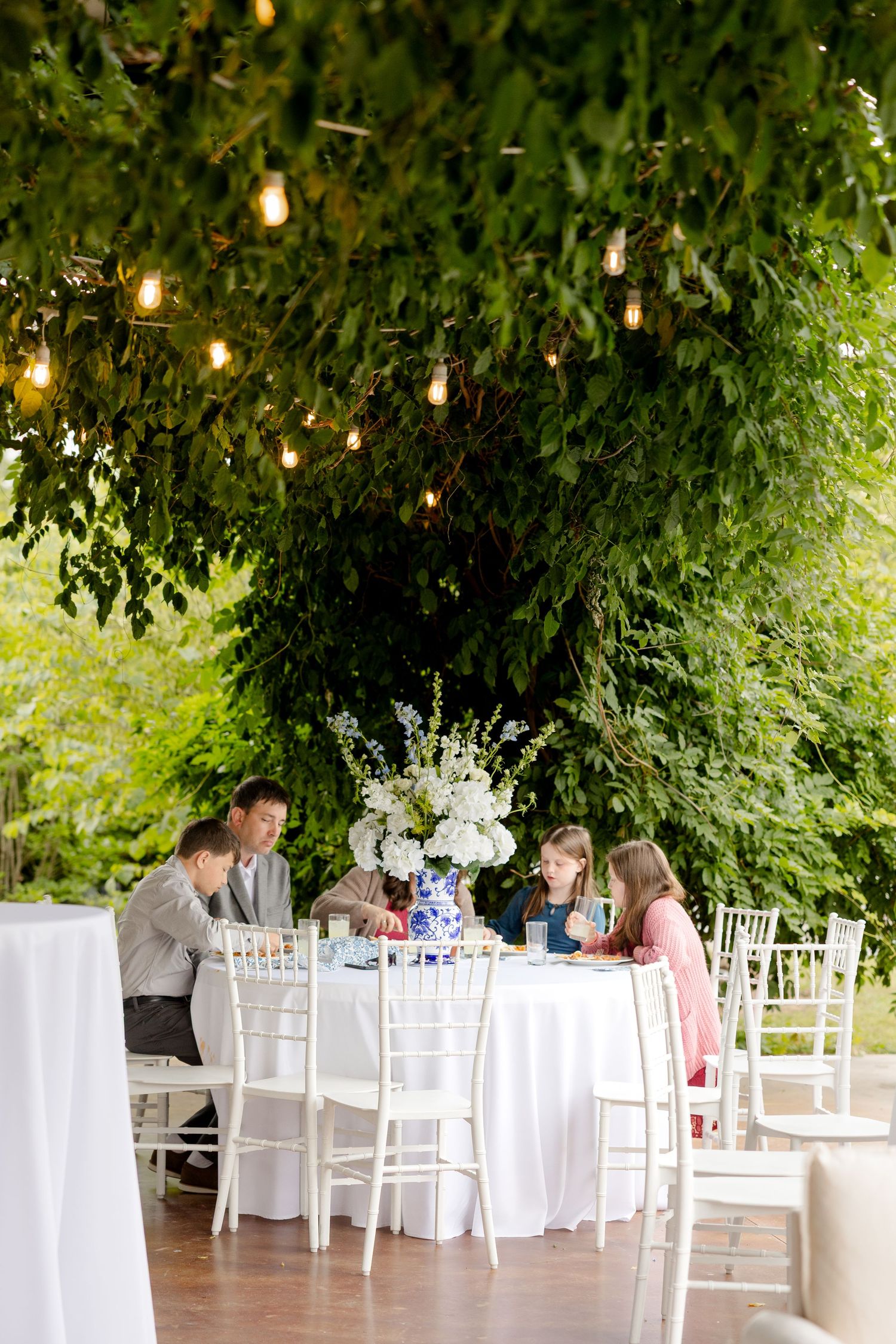 Outdoor wedding reception area with string lights and white tables under leafy green canopy.Cold Springs Events