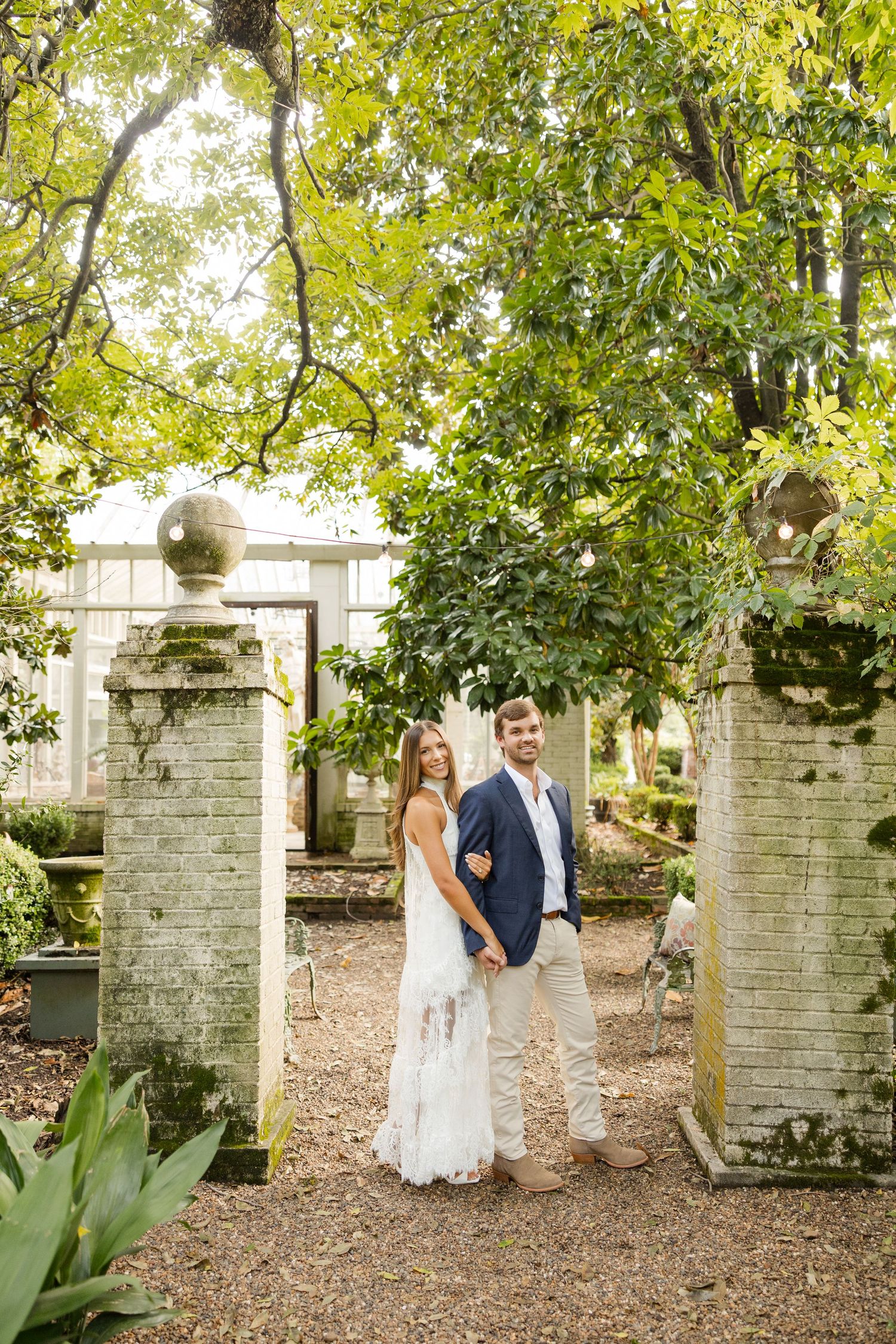A couple takes engagement photos among stone pillars and greenery in an outdoor garden setting.