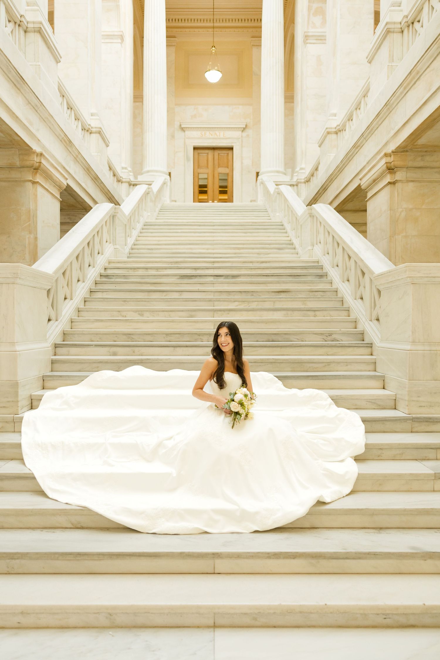 A white bridal gown spreads gracefully across grand marble steps in a classic architectural setting.