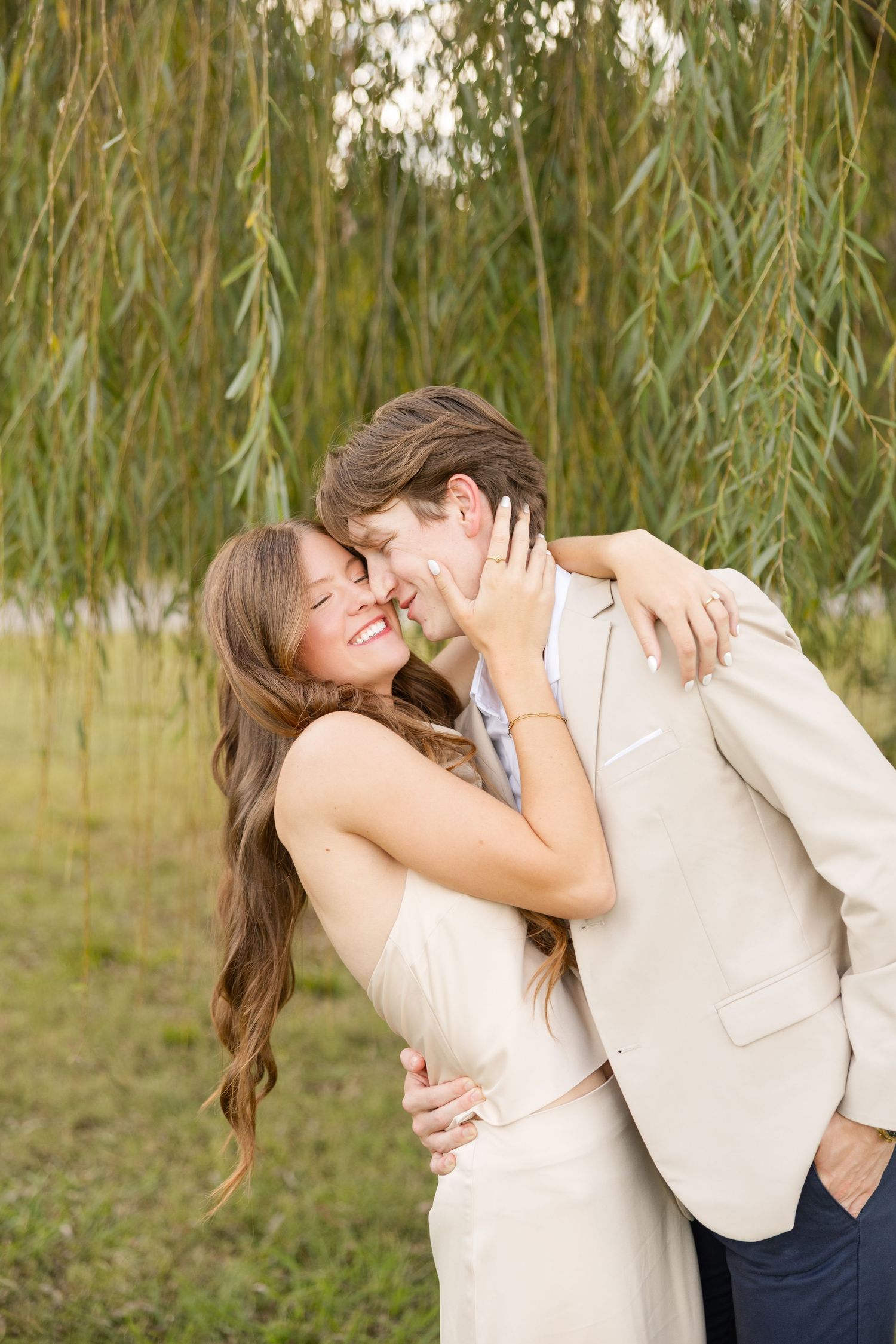 A romantic couple in light colored clothing share intimate moments among willow trees in a natural outdoor setting.