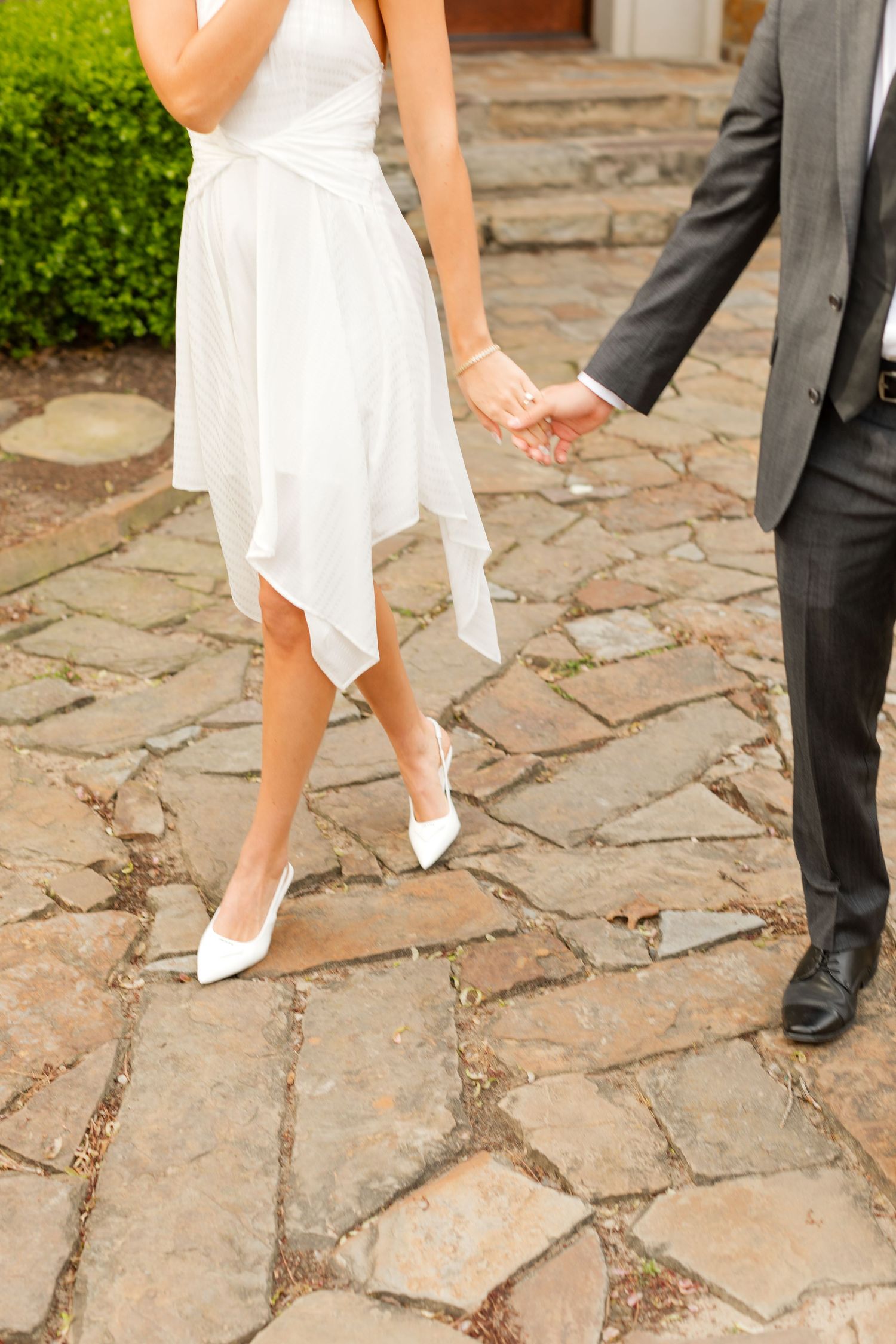 A couple in formal attire stands on a stone patio, with white shoes and dress visible from the knees down. Goodwin Manor.