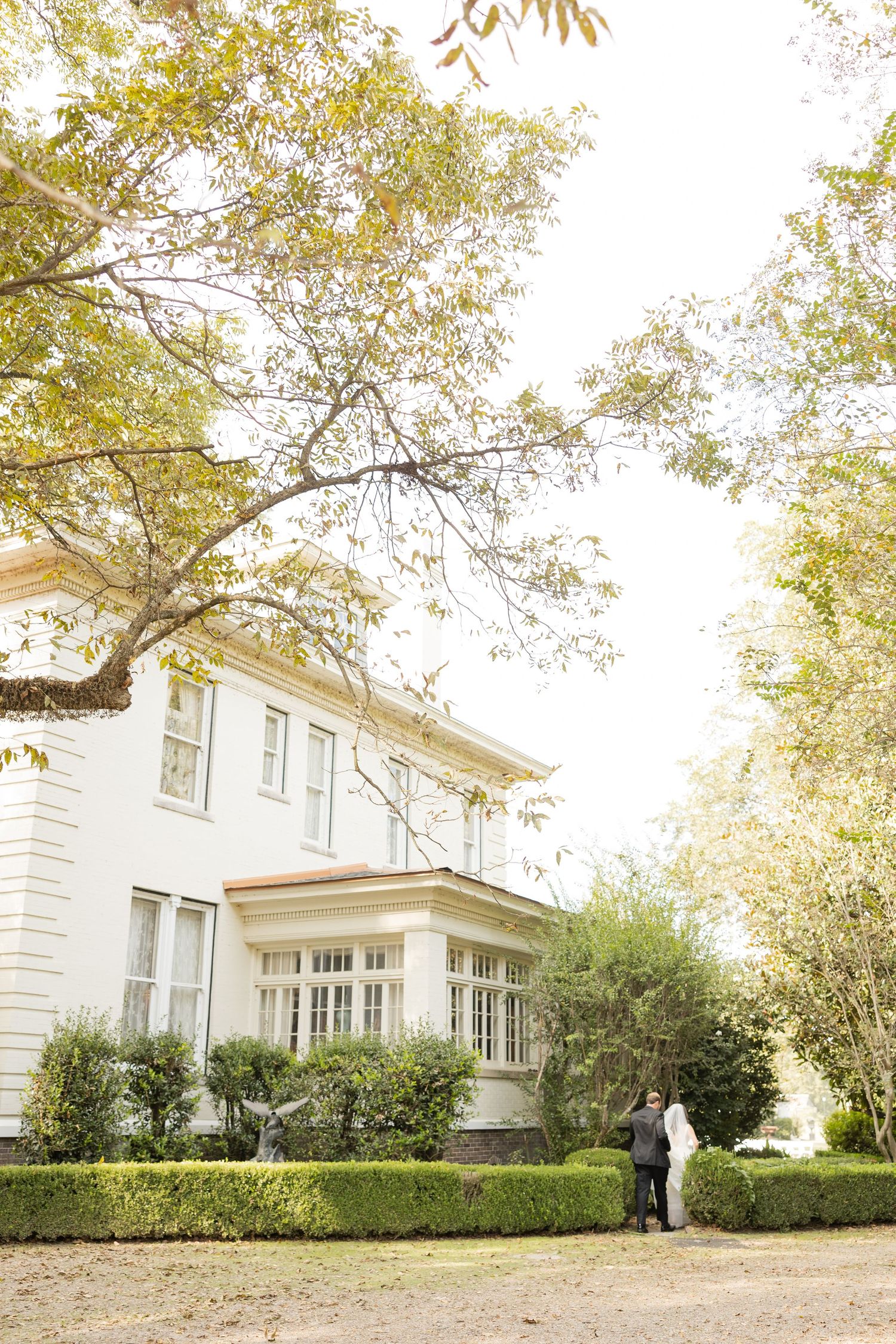 A panoramic view of a white colonial-style house with manicured hedges and trees in the front yard.