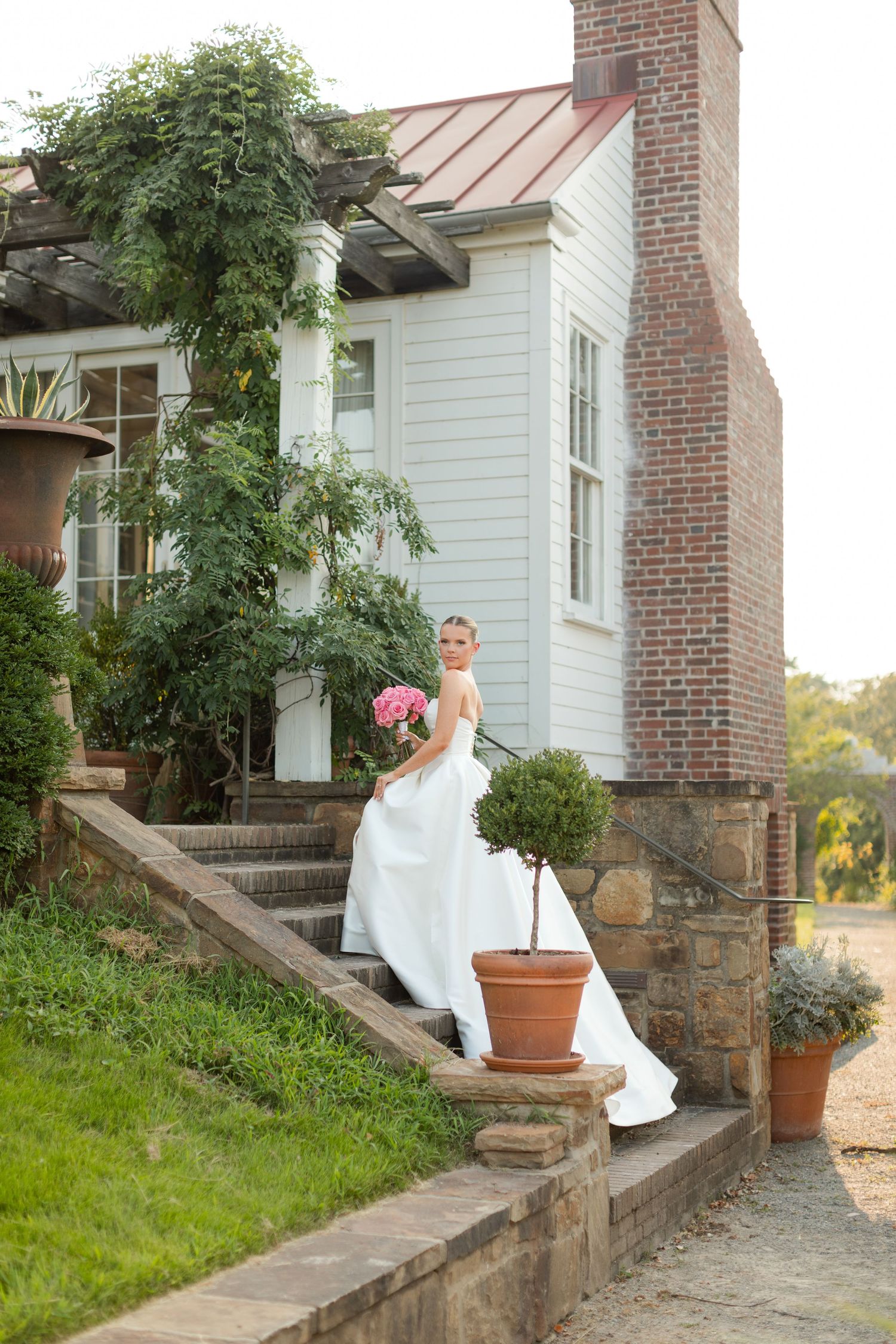 White wedding dress flows elegantly against rustic brick and stone steps of a charming farmhouse at sunset.