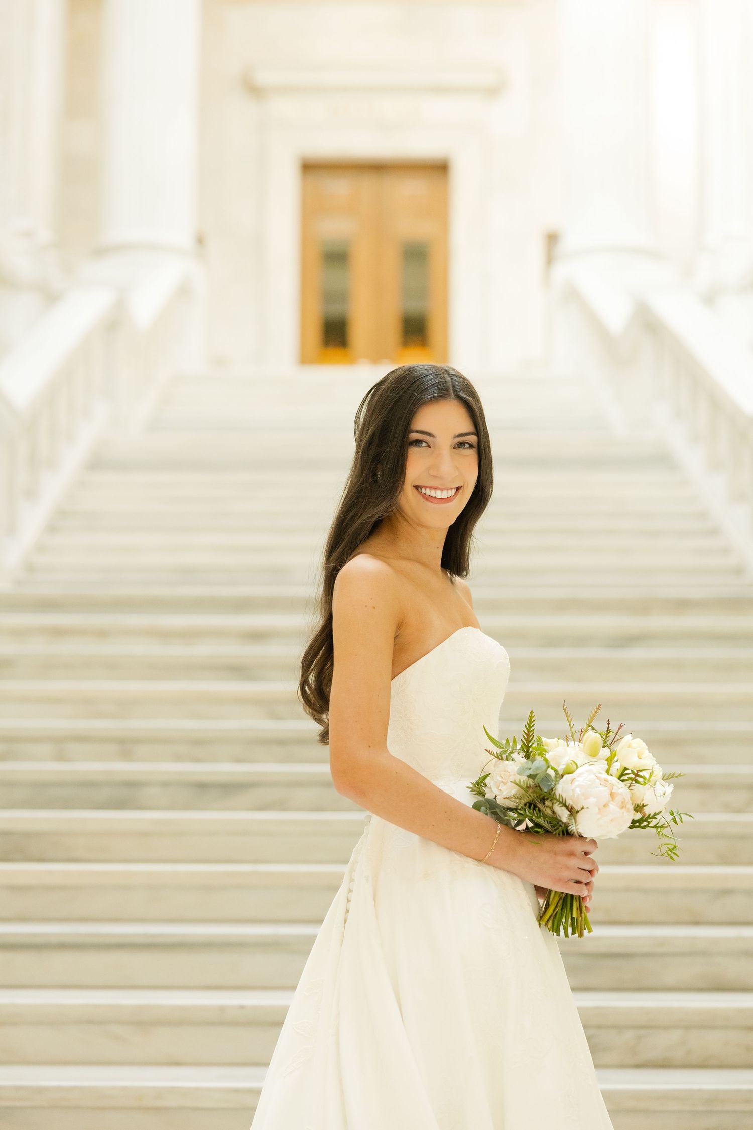 A bride in a white strapless gown holds a delicate bouquet of white flowers on grand marble steps.