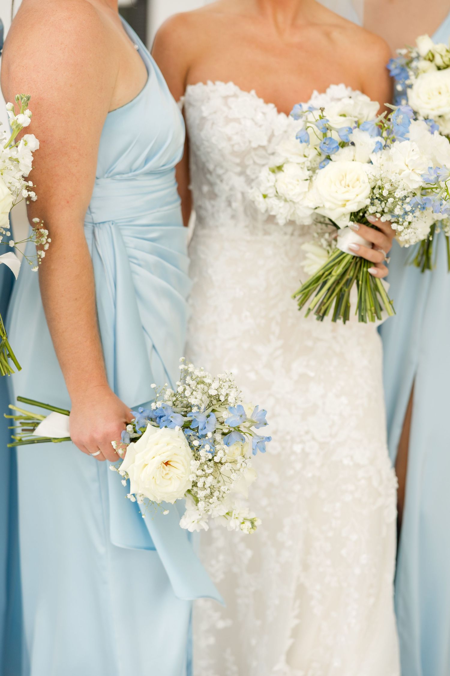 Bridesmaids in light blue dresses hold white floral bouquets next to bride in white lace gown.