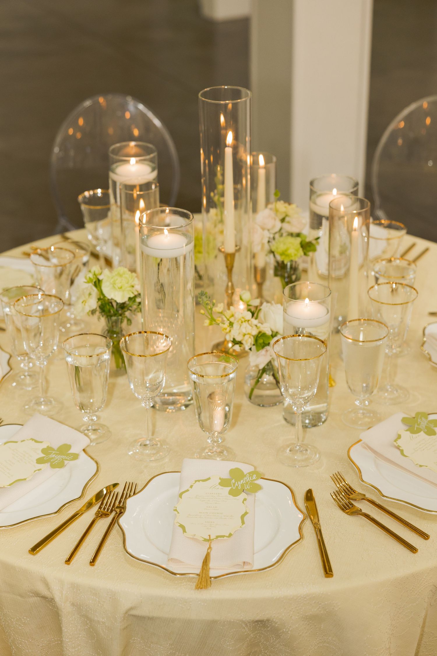 Elegant wedding reception table with gold cutlery, white plates, candles, and floral centerpieces on cream tablecloth.