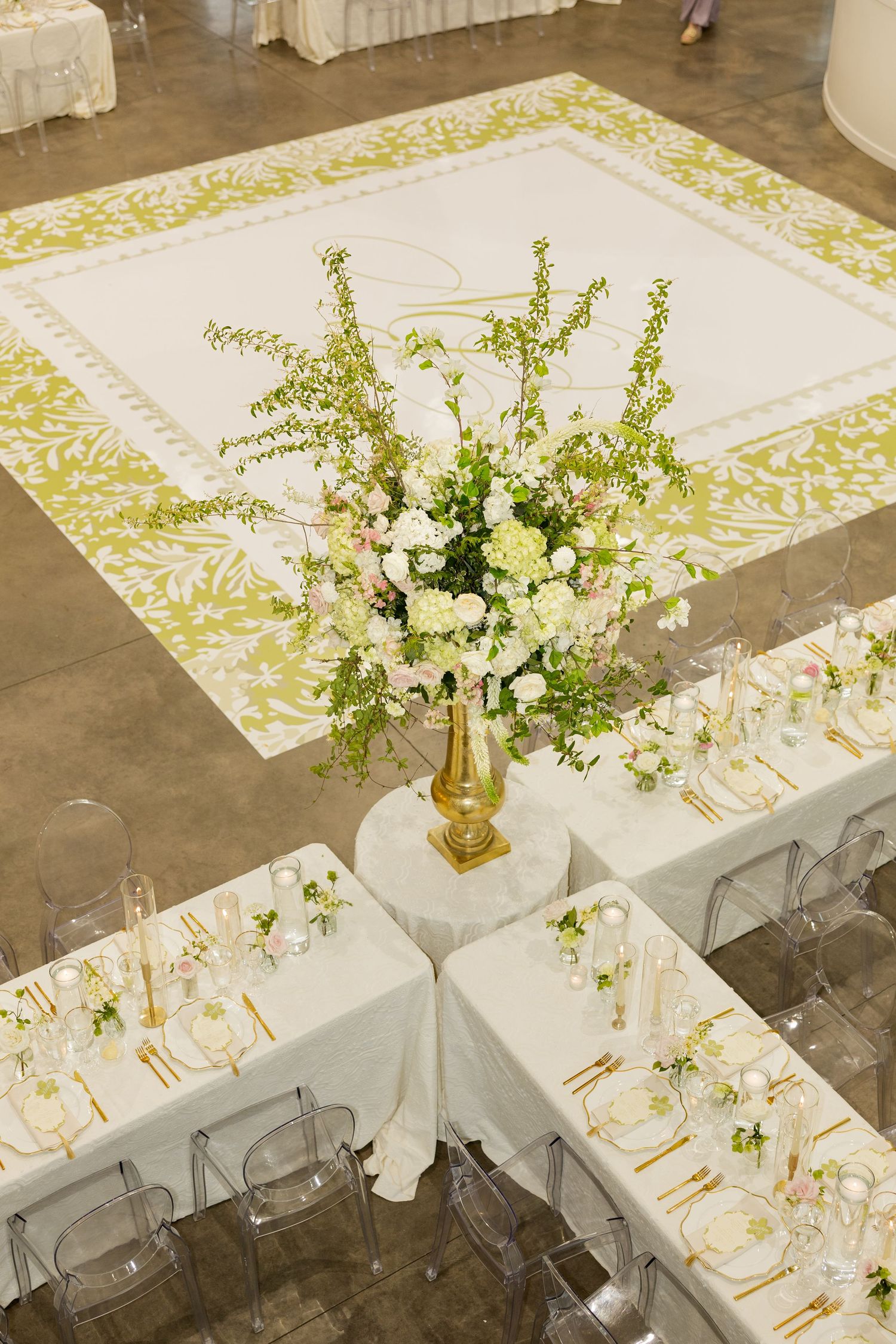 Luxurious wedding reception with white tables, ghost chairs, gold floral centerpiece, and ornate yellow dance floor.