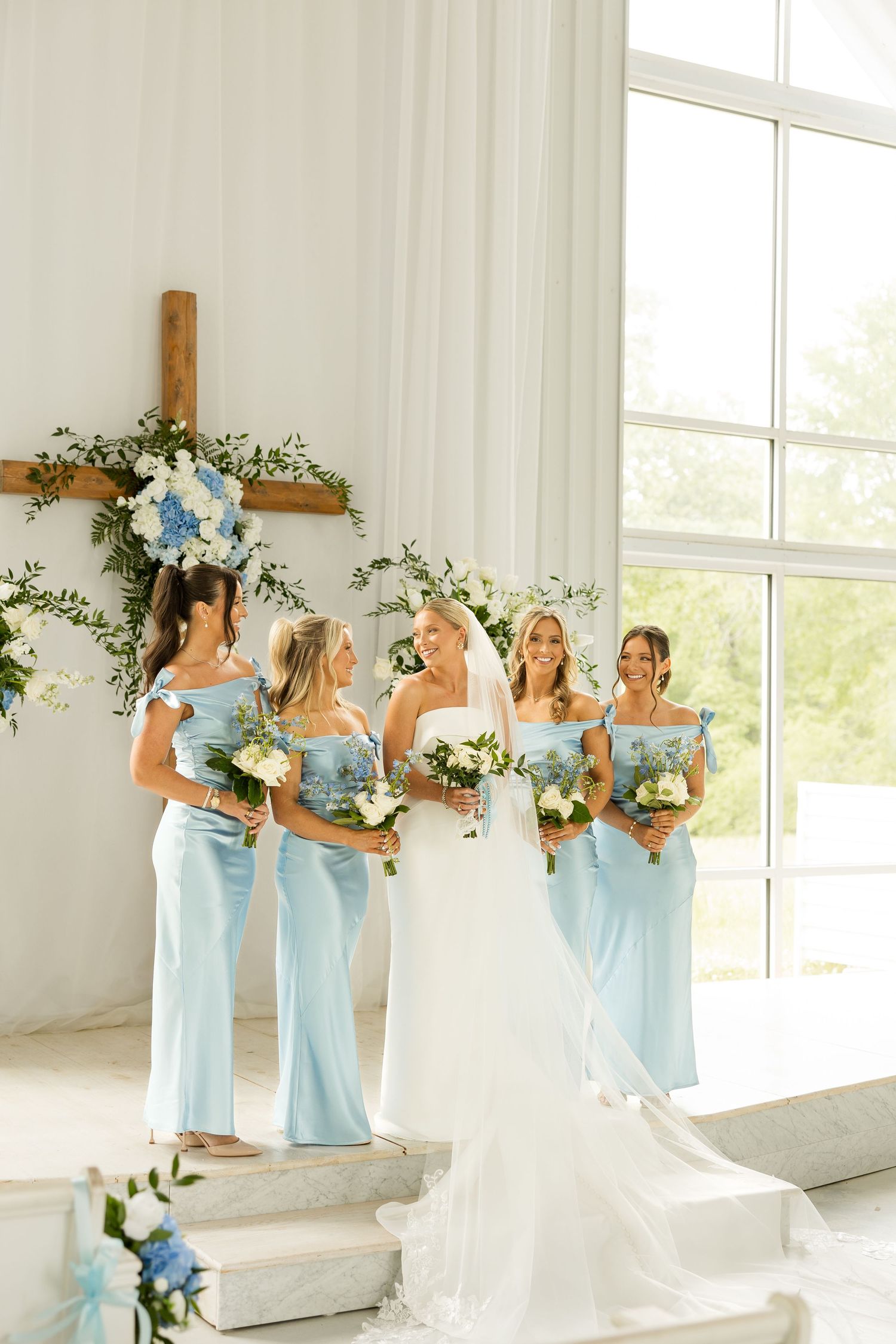 Bridesmaids in light blue dresses pose with bouquets by a window in a white chapel. Cold Springs Events.