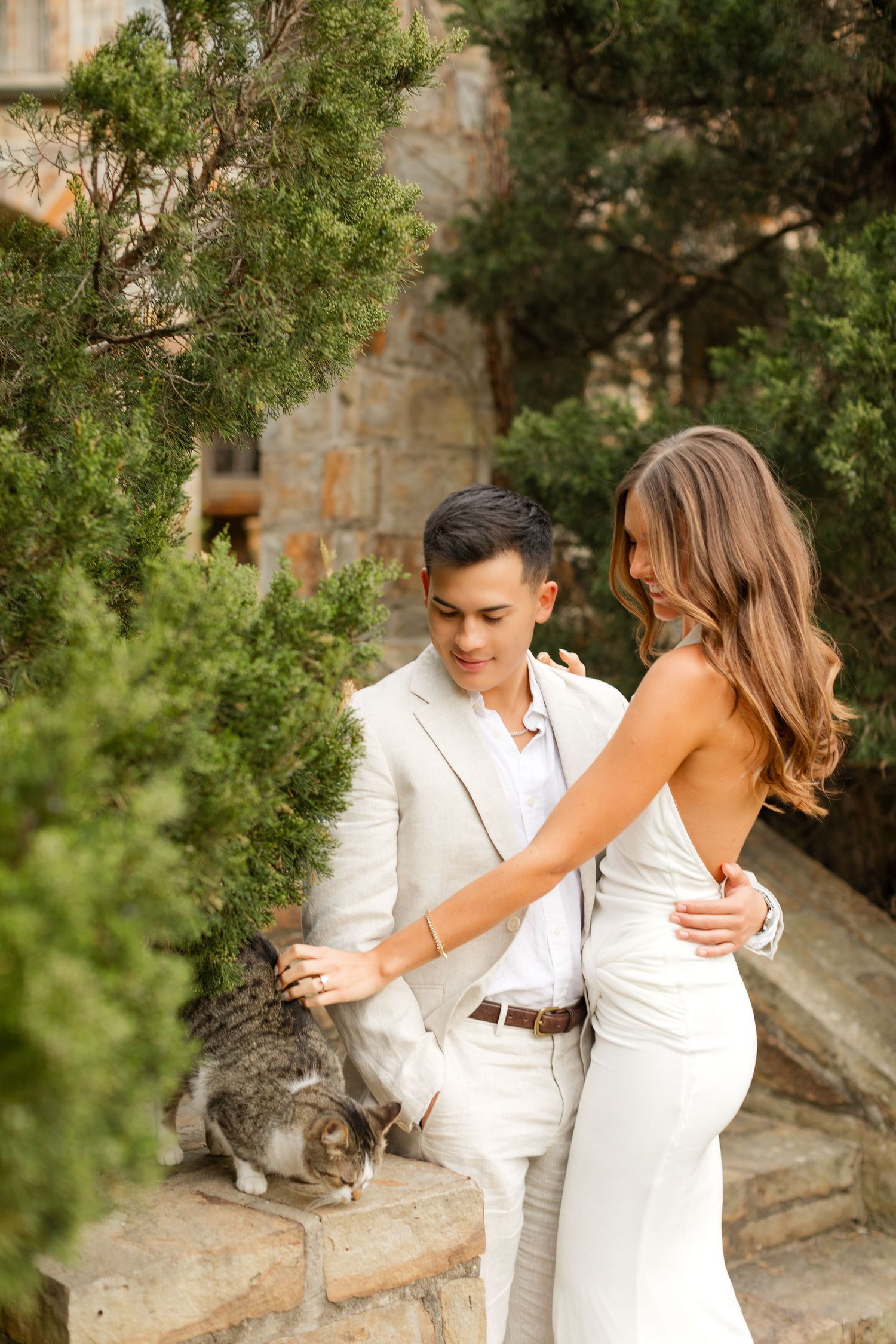 A couple in matching white outfits poses together near stone steps with a cat lounging in the background. Goodwin Manor.