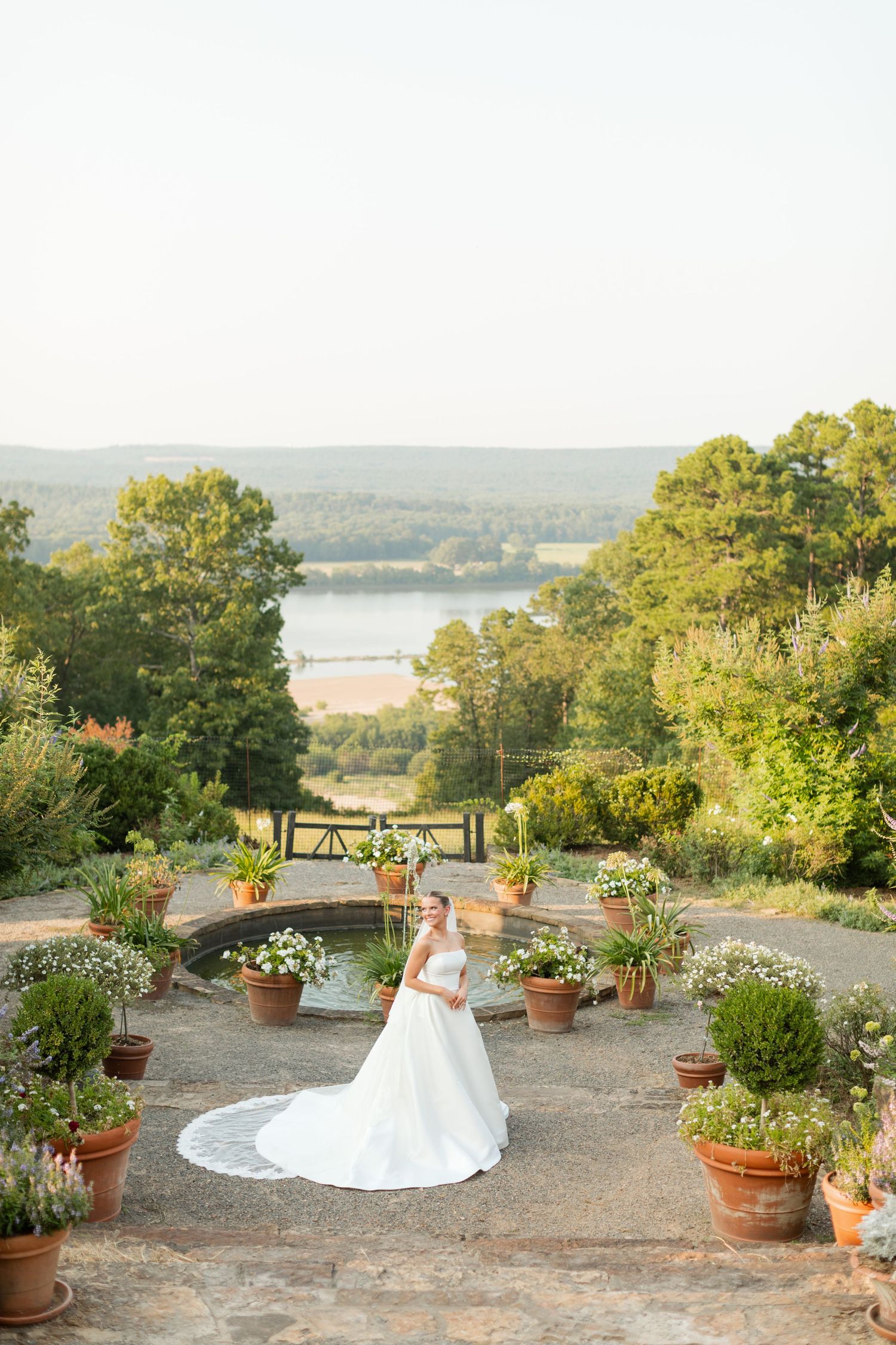 A scenic outdoor terrace with potted flowers overlooking a lake and rolling hills during golden hour.