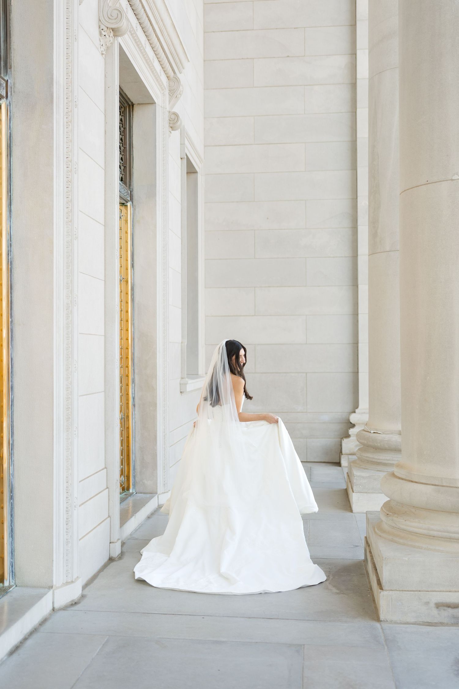A bride in a flowing white dress stands gracefully between classical marble columns.