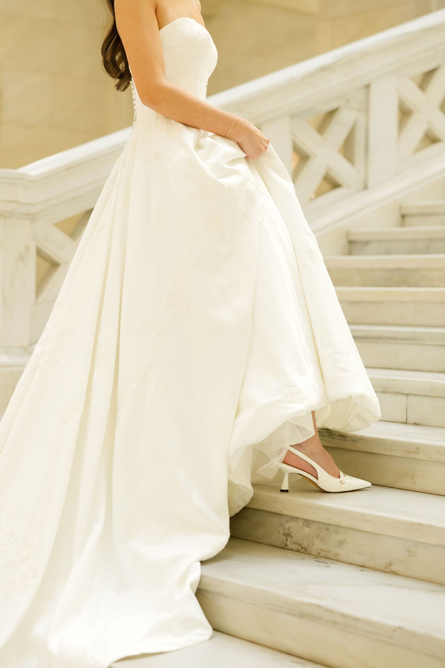 A bride in a flowing white wedding dress sits elegantly on marble stairs, showing off her white heels.