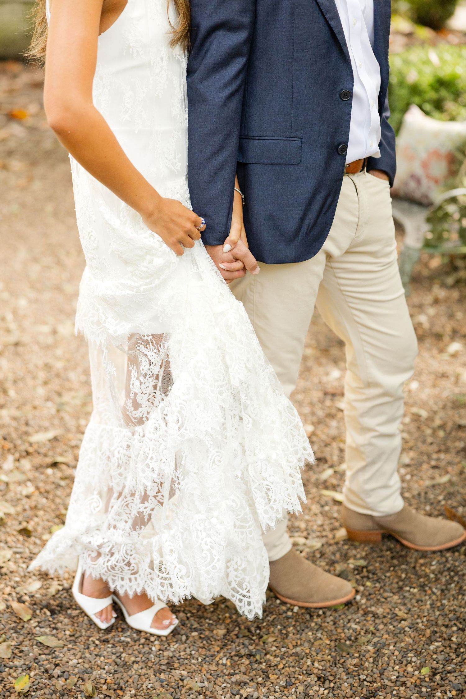 Close-up of wedding attire showing a white lace dress hem and navy suit jacket with khaki pants on a gravel path.
