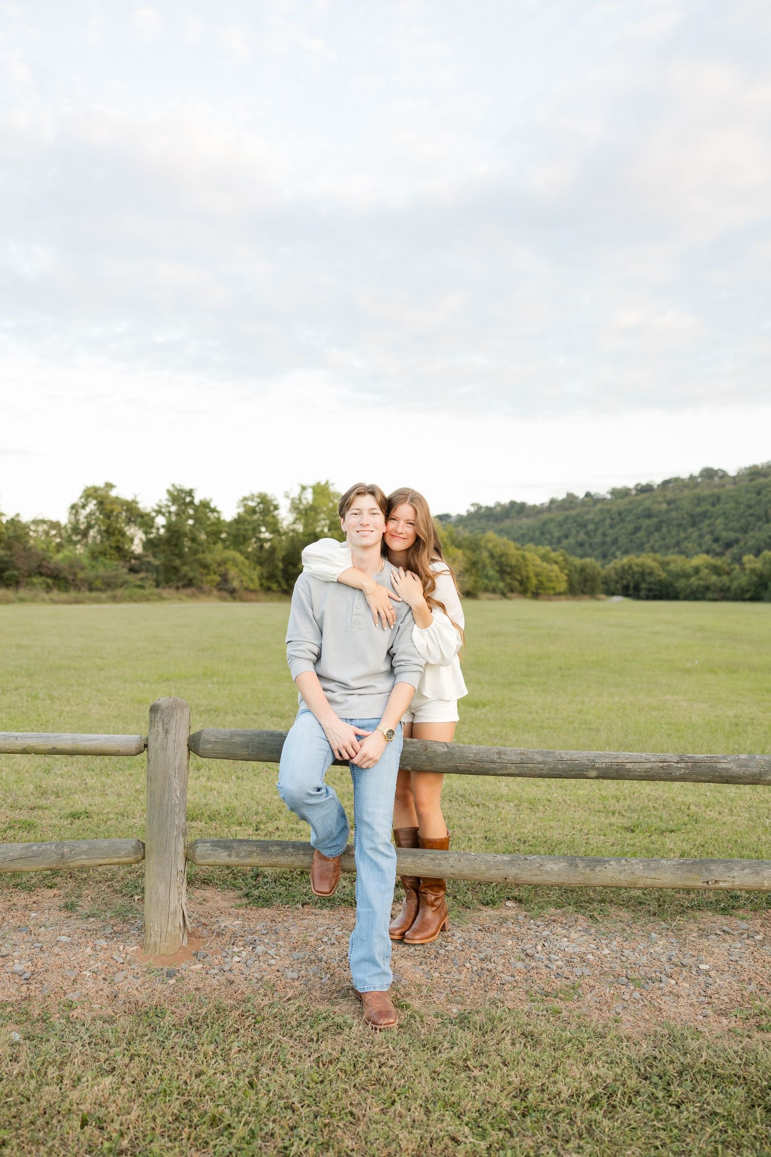 A couple shares a tender moment while sitting on a wooden fence in a pastoral field setting.