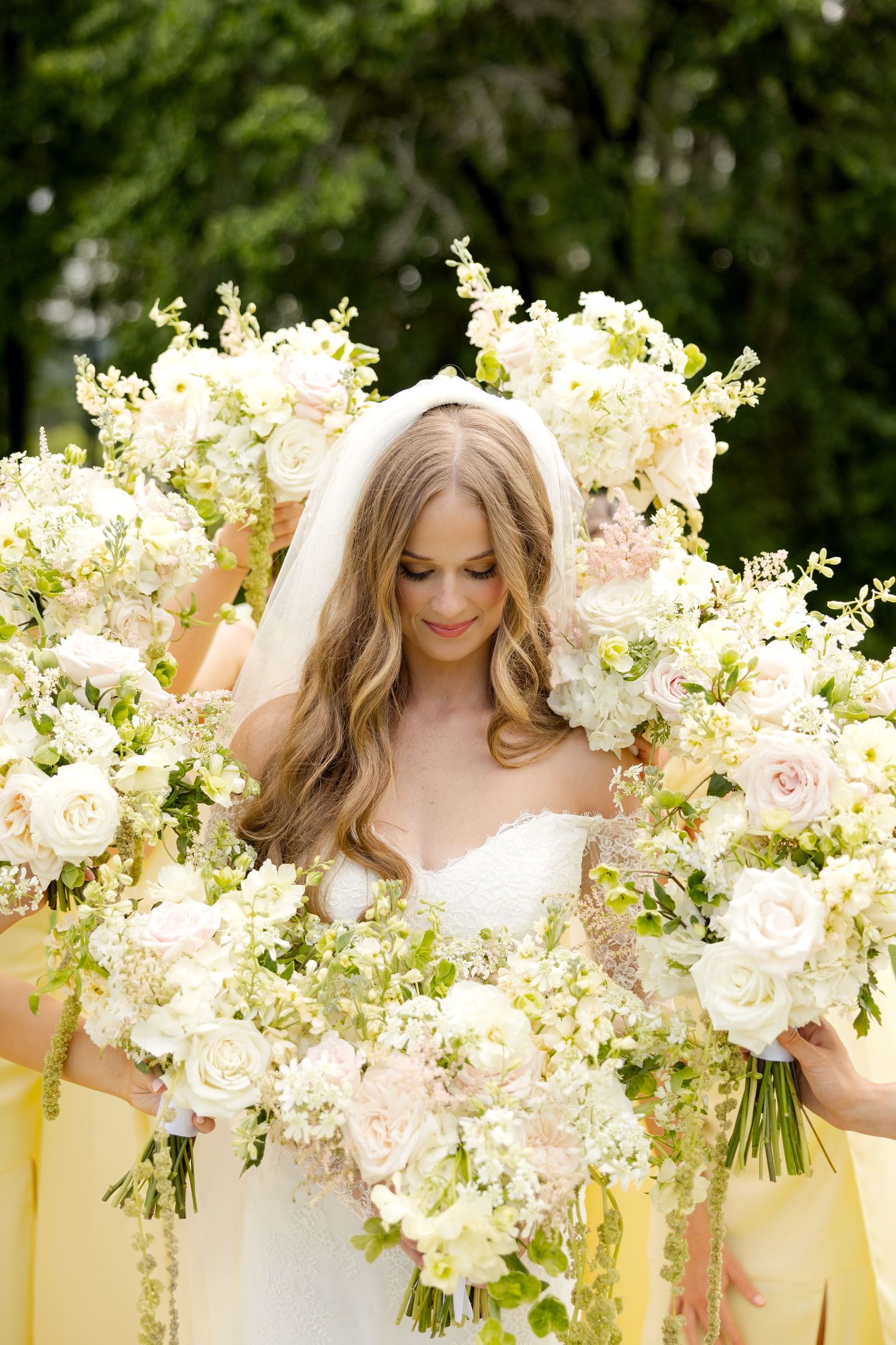 Bride in white gown and veil surrounded by bridesmaids holding large white and blush floral bouquets outdoors.