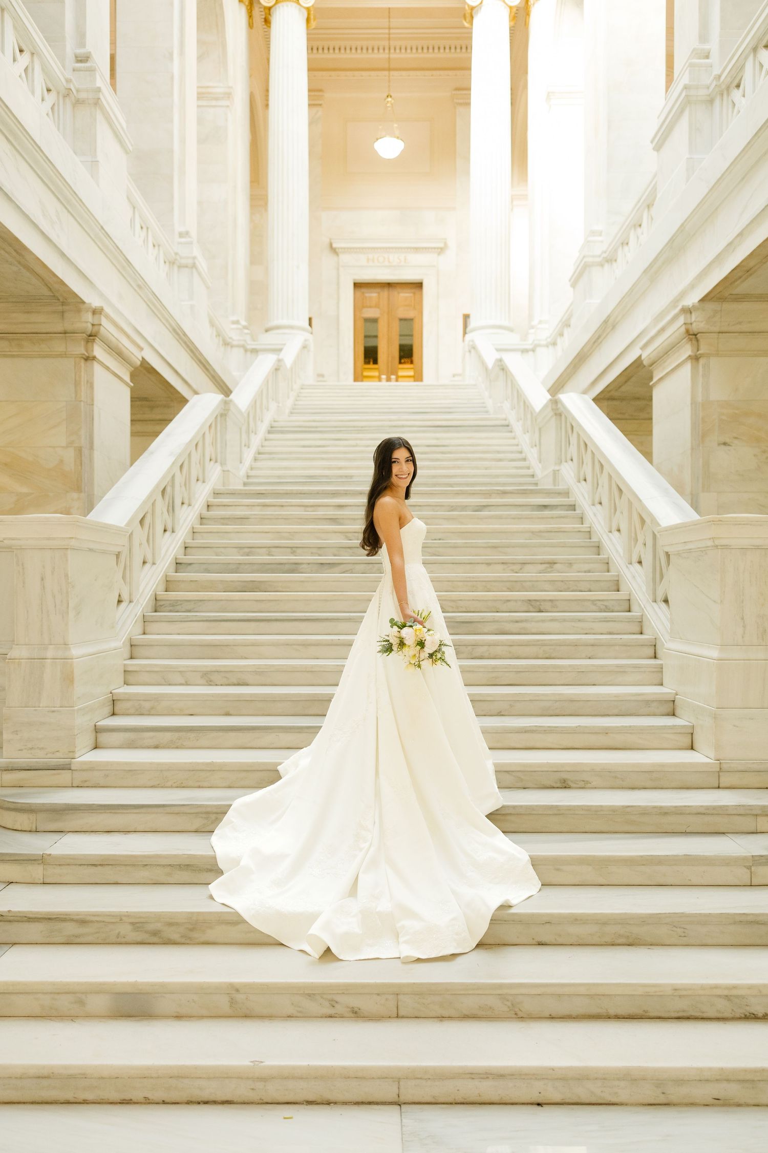 A bride in a flowing white wedding gown stands on elegant marble stairs in a grand historic building interior.