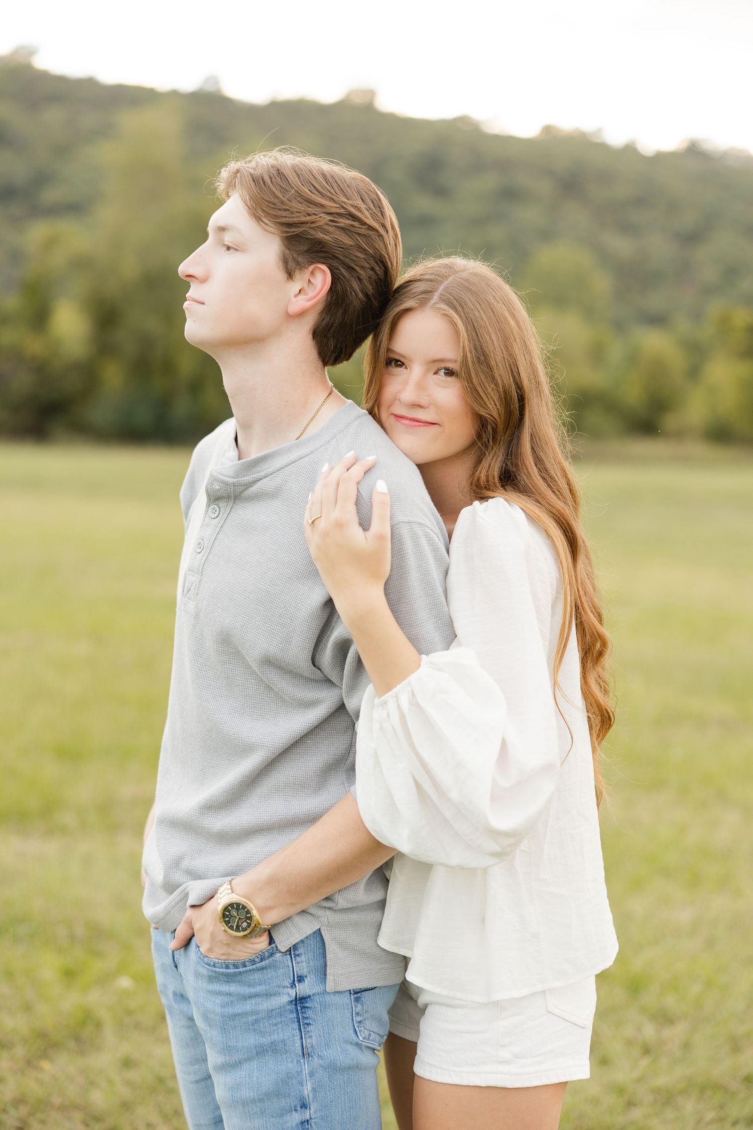 A couple embraces in a grassy field during golden hour, with her in white and him in gray casual wear.