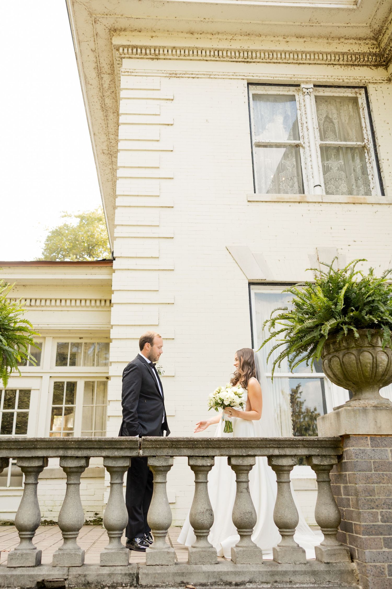 A couple stands on a white balustrade terrace with hanging plants and decorative columns of a historic building.