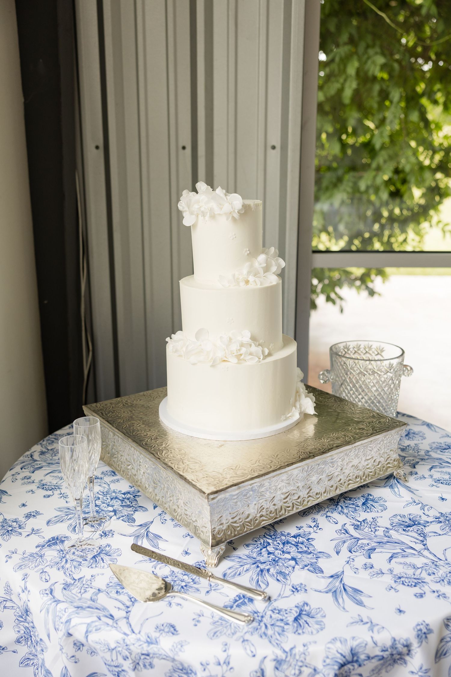 Elegant three-tier white wedding cake with floral decorations displayed on a silver stand with blue floral tablecloth.Cold Springs Events