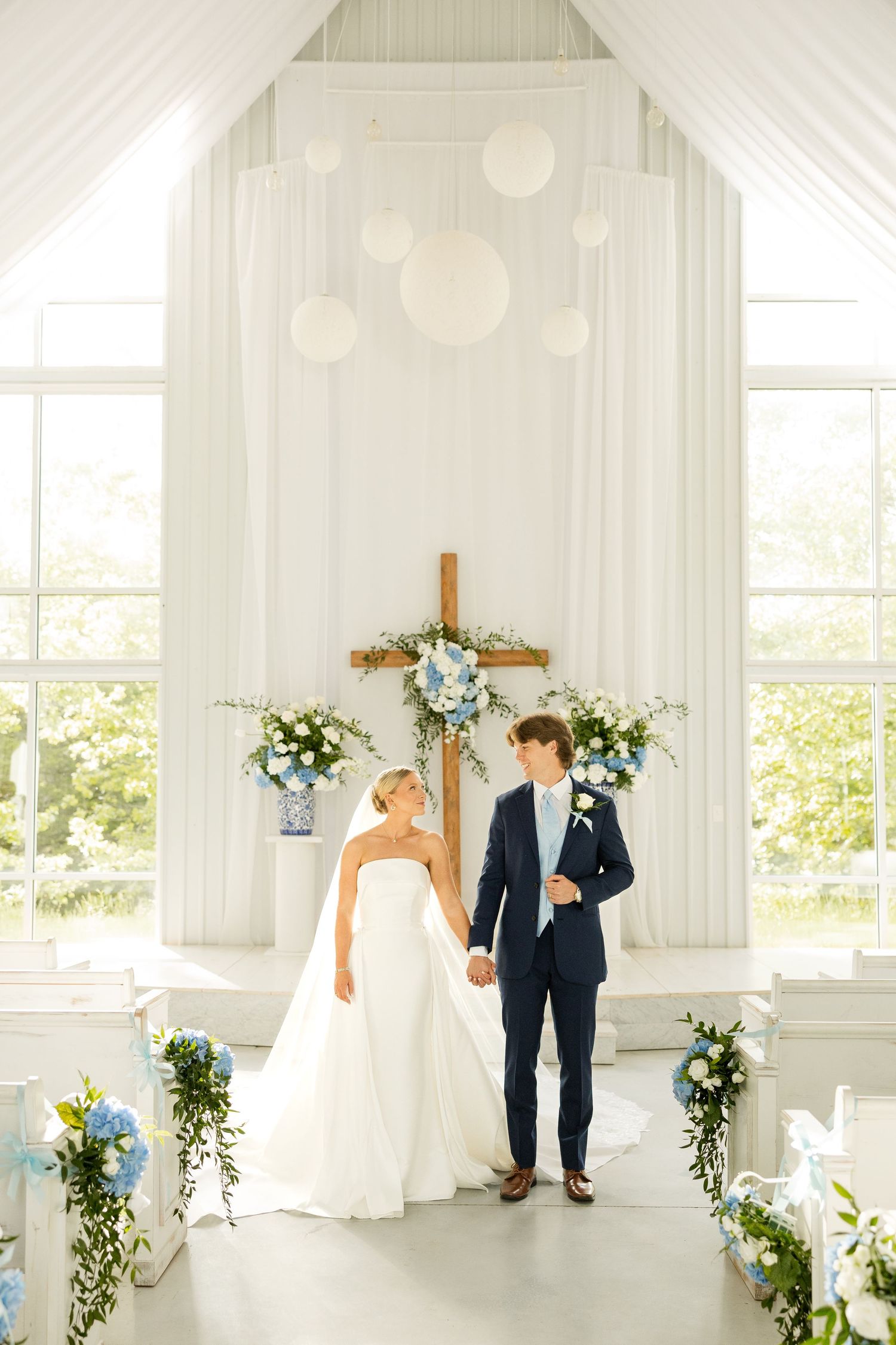 Wedding couple walking through a sunlit white chapel decorated with blue and white floral arrangements.Cold Springs Events