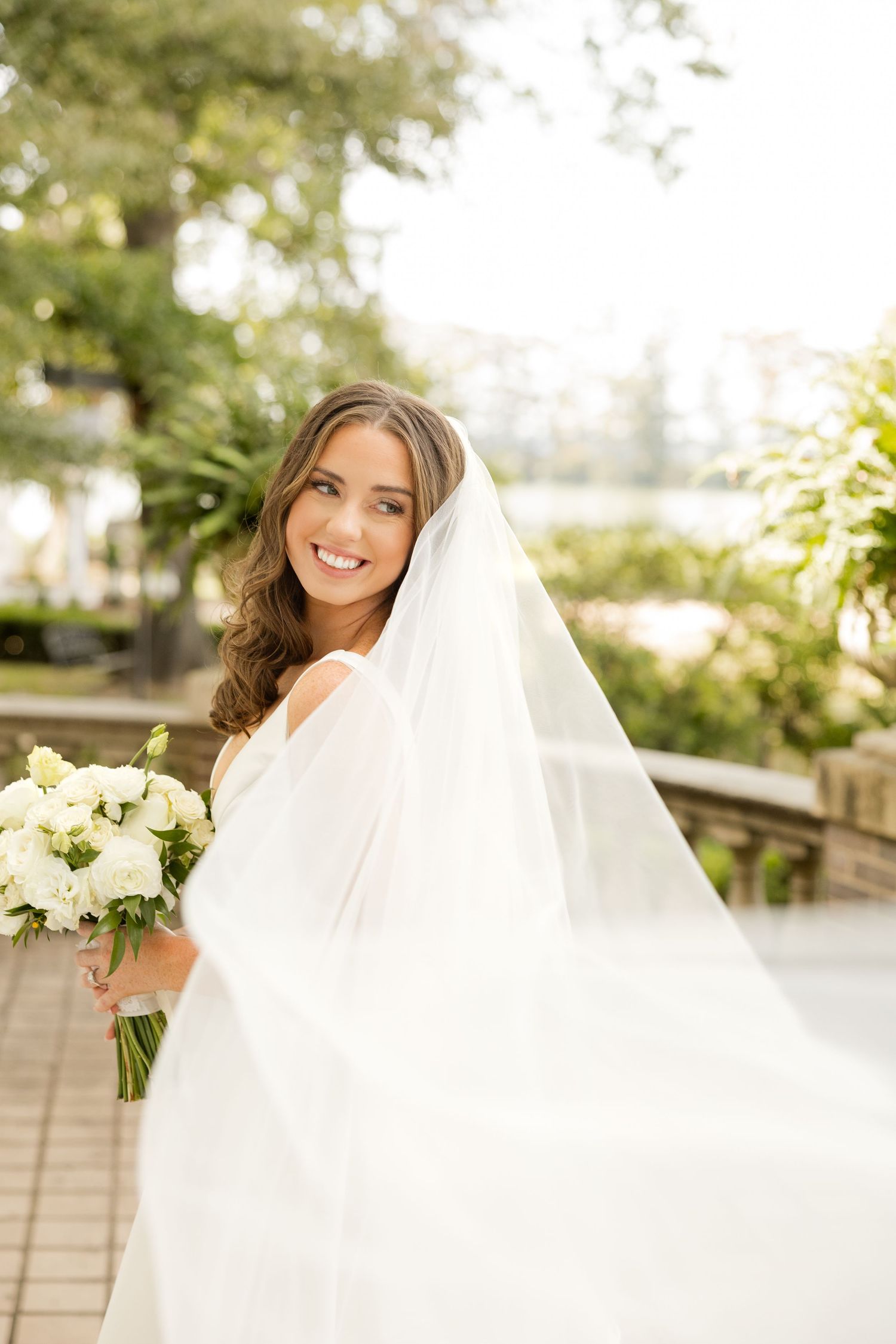 Bride in white dress holding bouquet outdoors with flowing veil in sunlight.Marlgsate Plantation