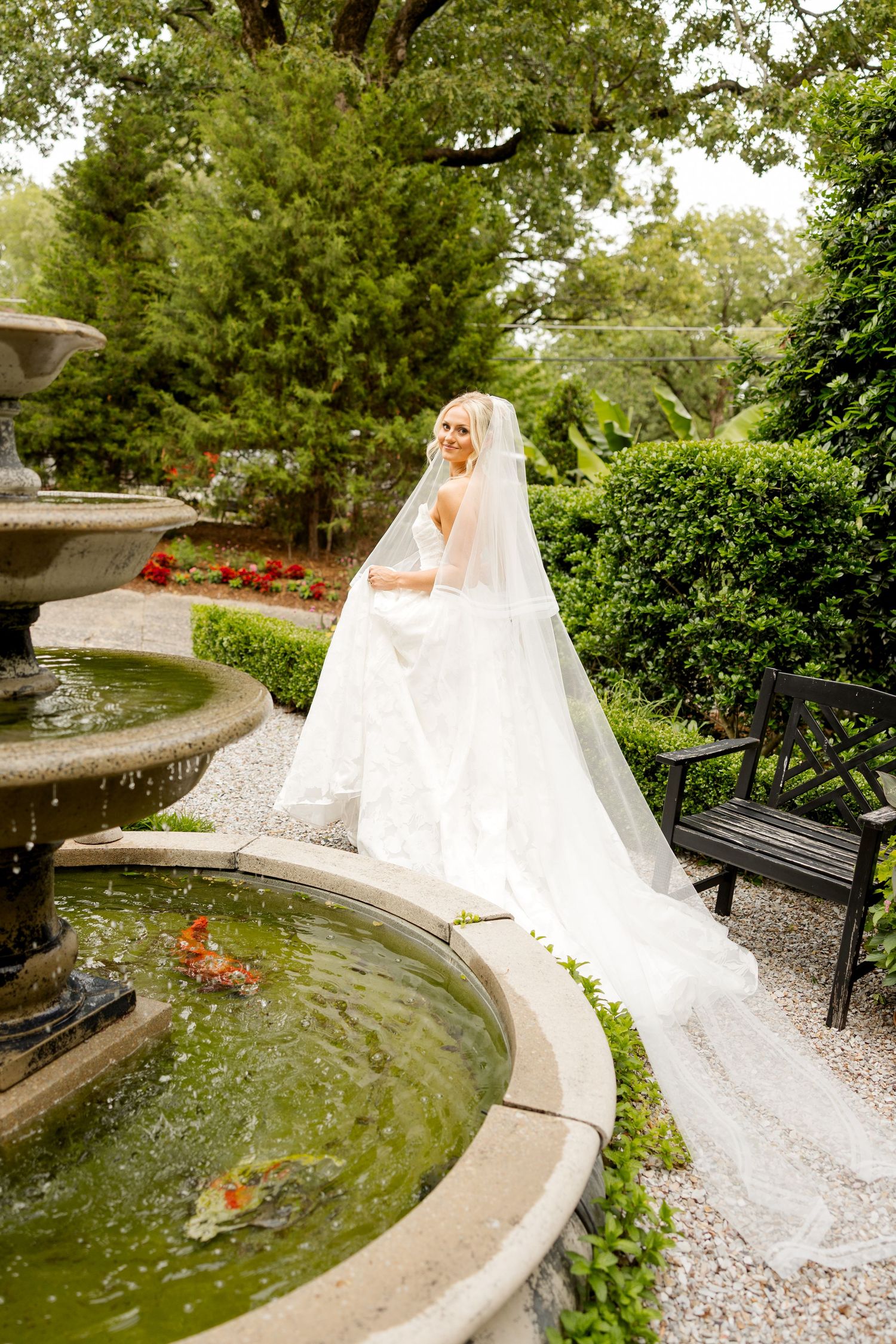 A flowing white wedding gown trails elegantly along a curved garden path beside a tranquil pond in a lush outdoor setting.