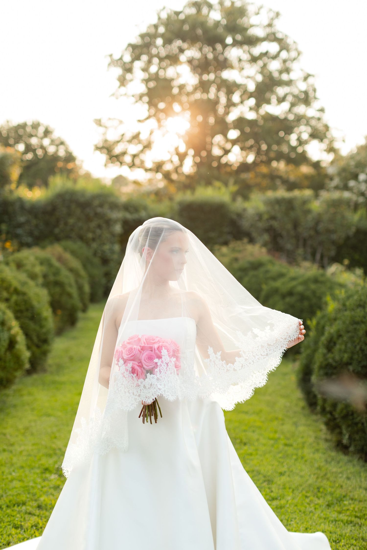 Classic white wedding gown with pink floral accents captured in a garden setting at sunset.
