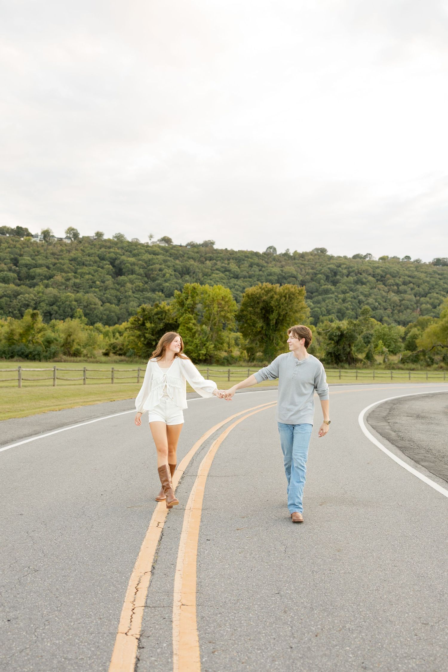 Two people hold hands while walking on opposite sides of a yellow road line with green hills in the background.