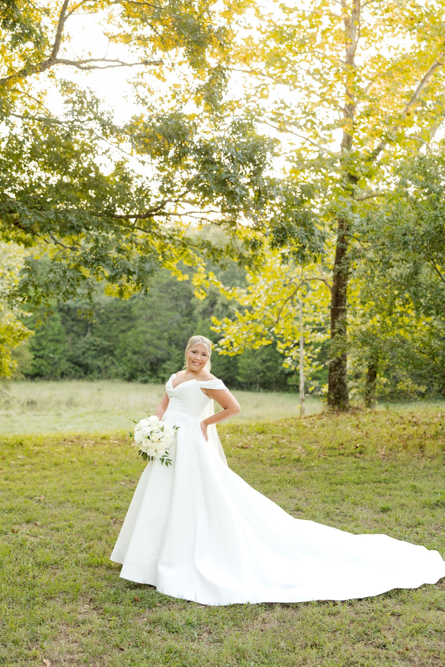 A flowing white wedding gown spreads across autumn grass as golden sunlight filters through trees in the background.