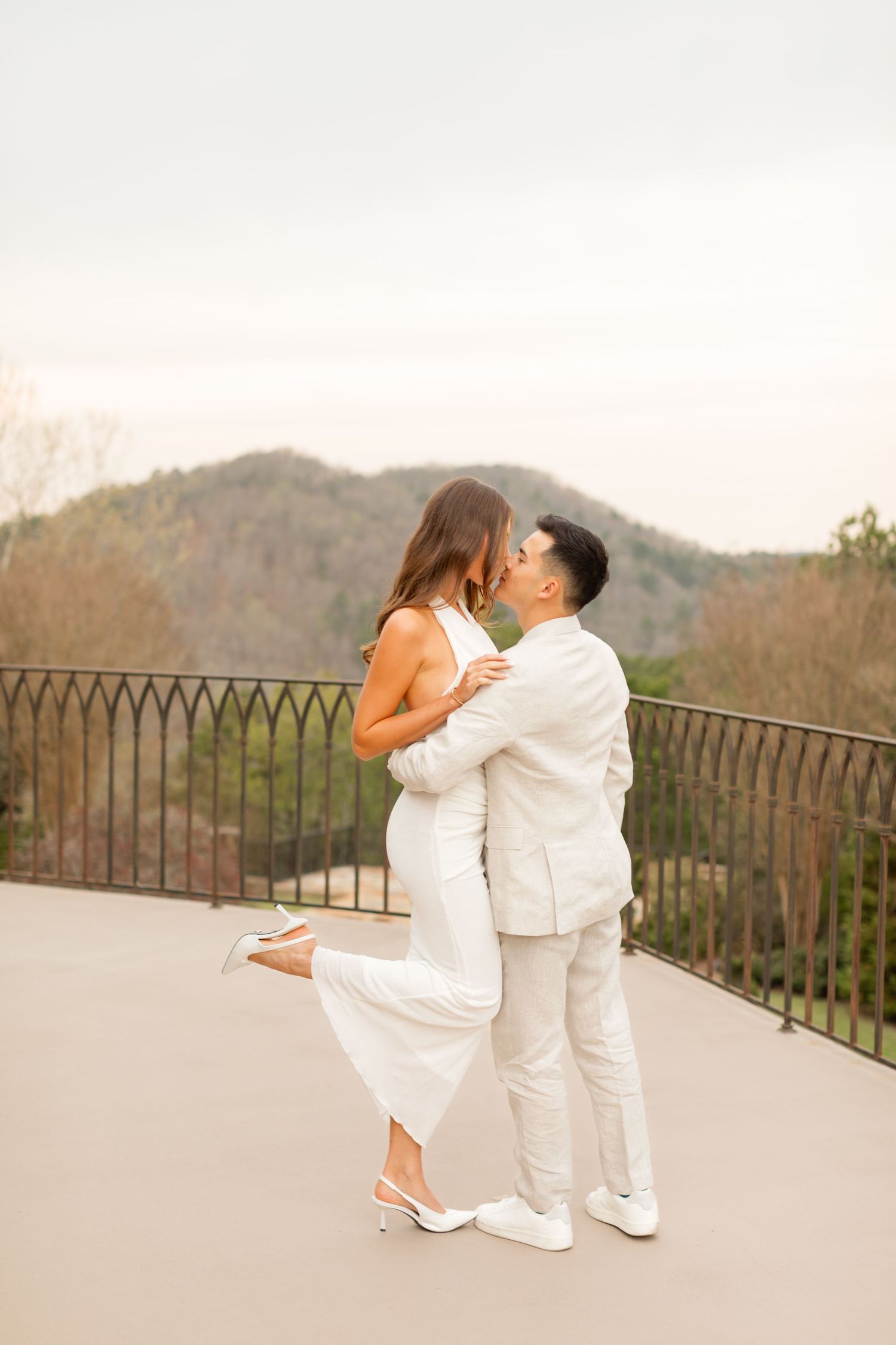 A romantic couple in white outfits sharing an intimate moment on a balcony overlooking mountain scenery at sunset. Goodwin Manor.