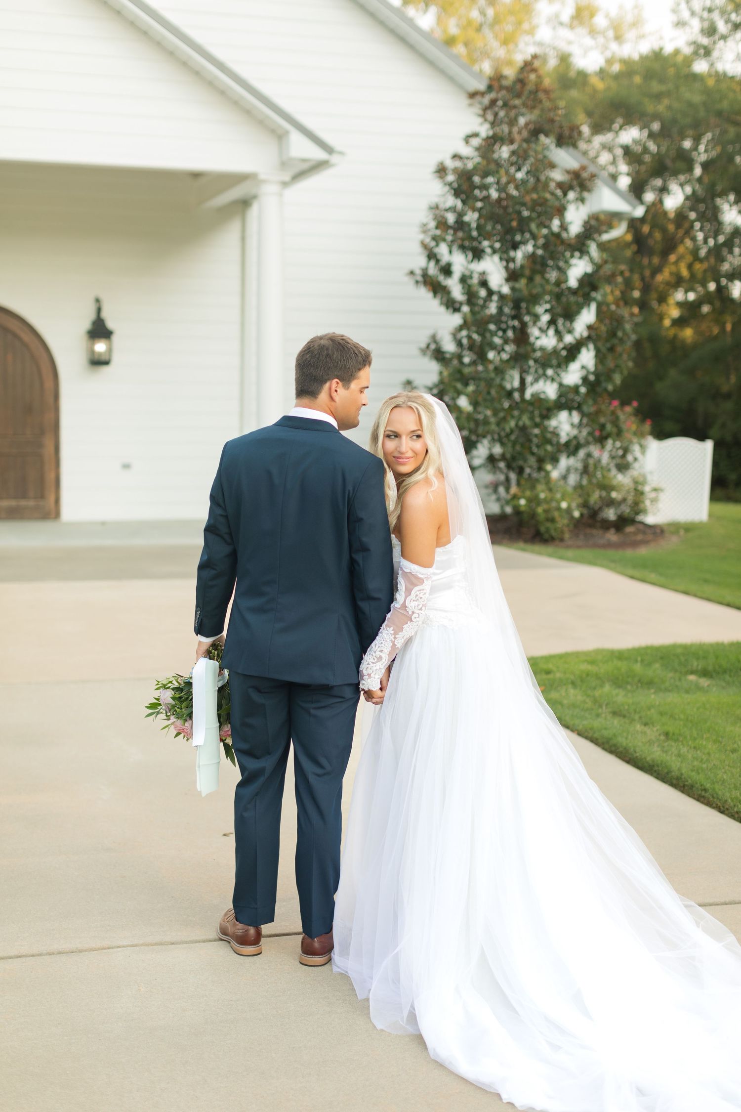 Couple walks along white church exterior in formal wedding attire. Legacy Acres.