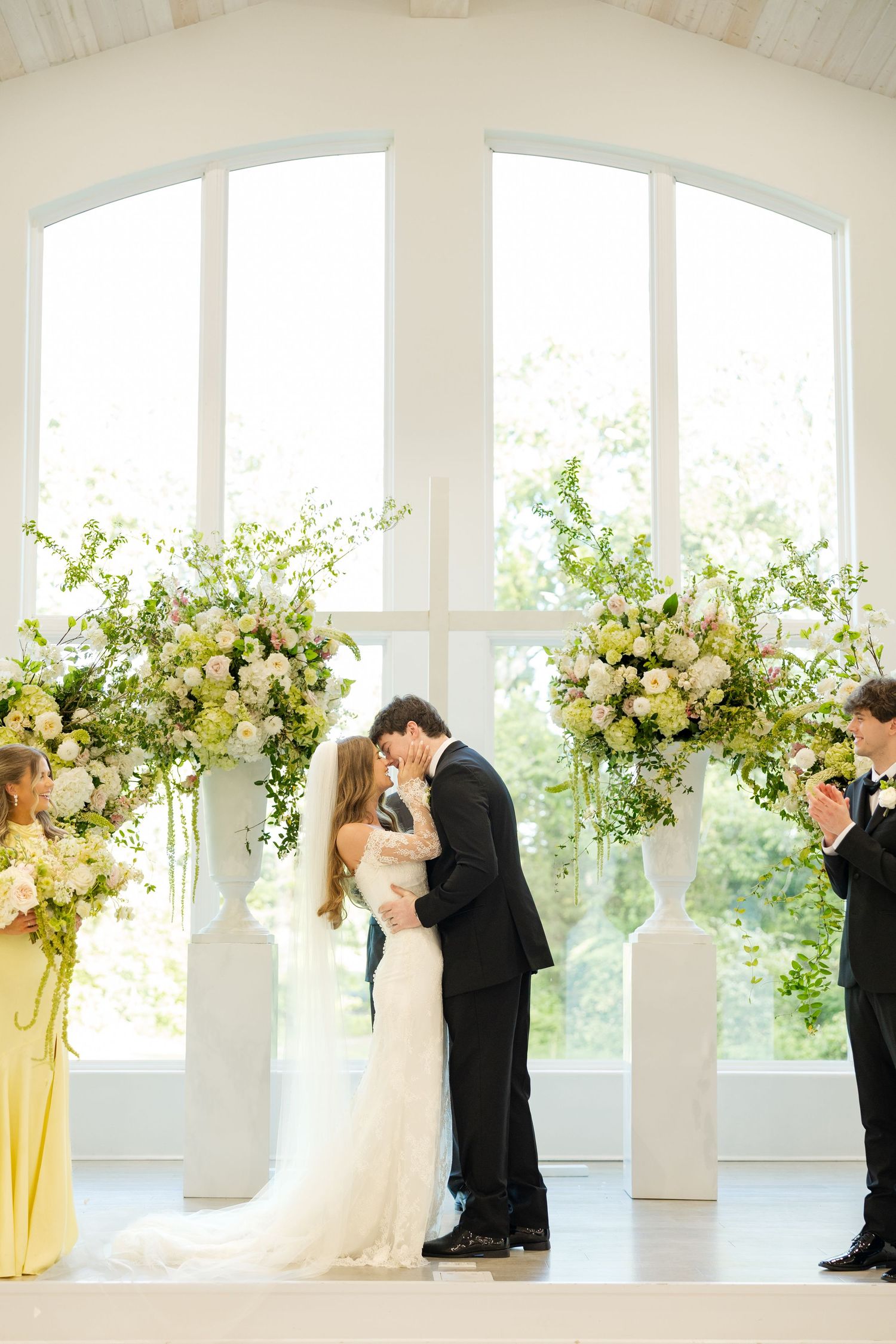 Bride and groom share first kiss at elegant wedding ceremony with tall white floral arrangements and arched windows.