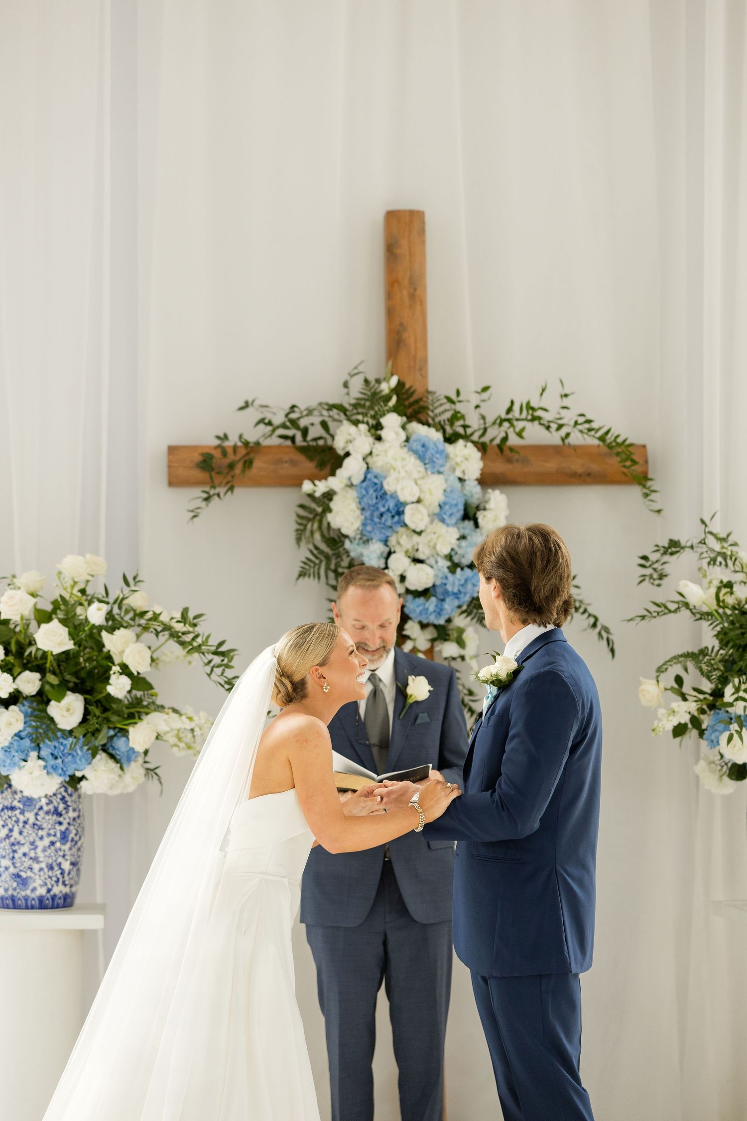 Wedding ceremony moment in white chapel with wooden cross and blue floral arrangements.Cold Springs Events