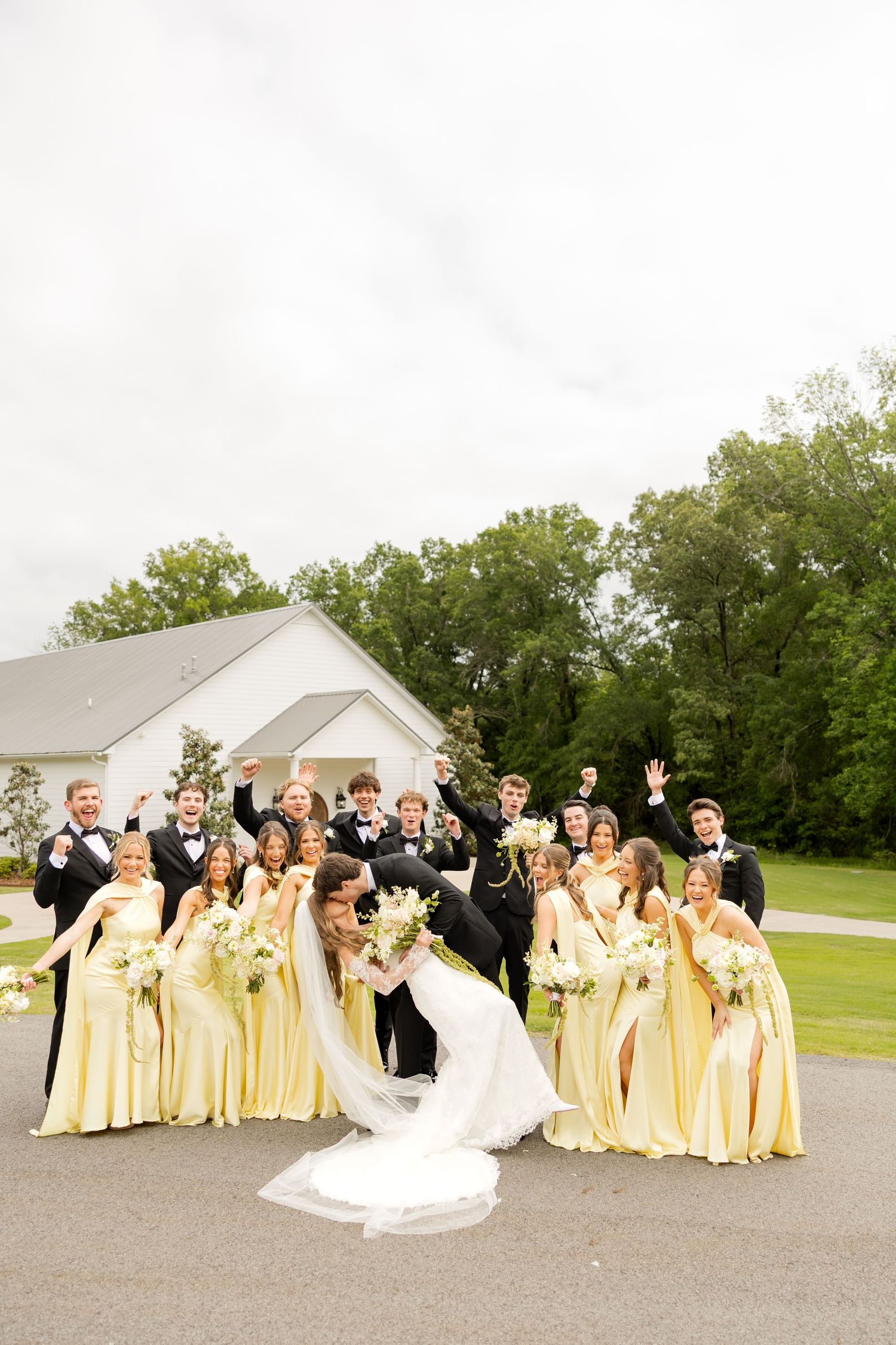 Wedding party celebrating outdoors, bridesmaids in yellow gowns and groomsmen in black tuxedos cheering as bride and groom kiss.