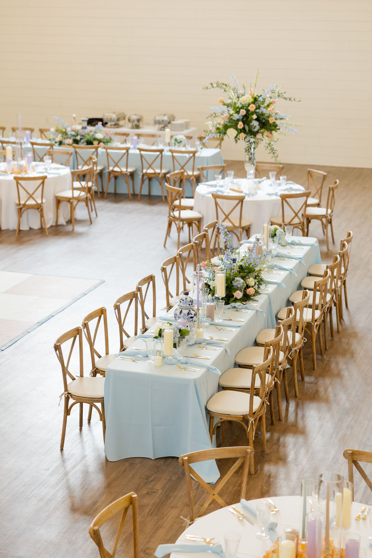 Long dining tables with light blue linens and wooden cross-back chairs create an elegant wedding reception setup.
