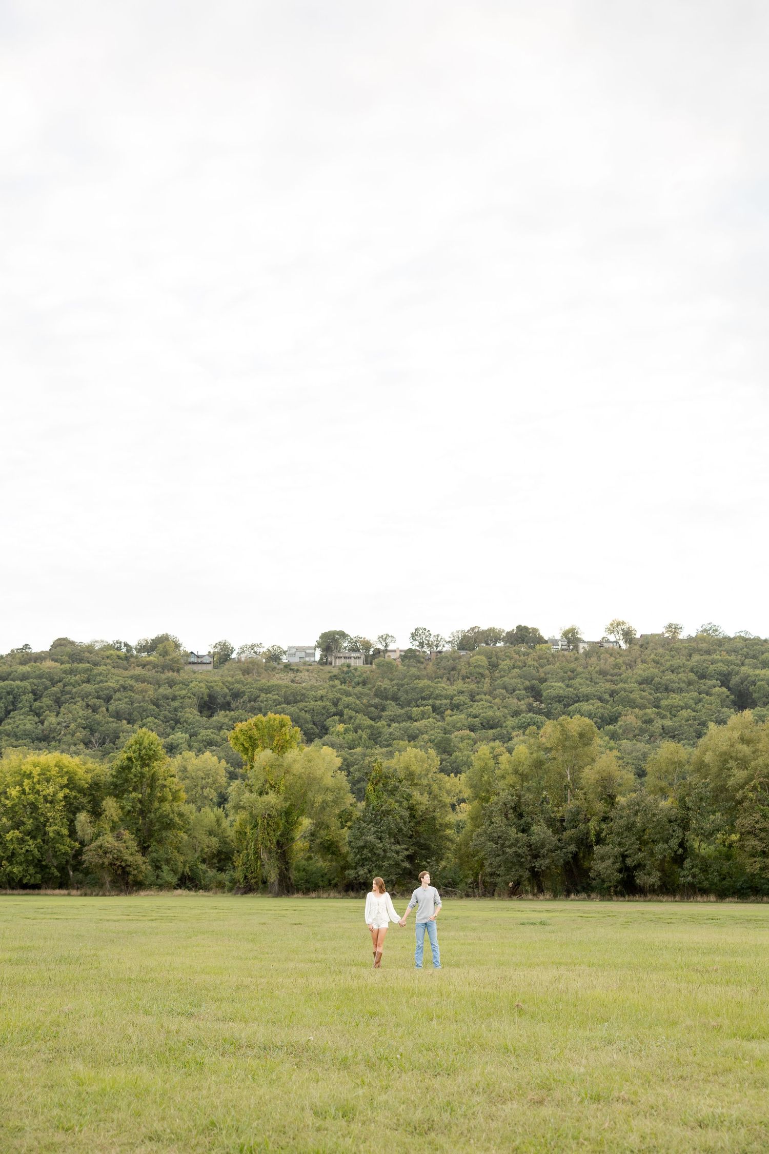 A couple walking together in a vast green meadow surrounded by trees on a sunny day.