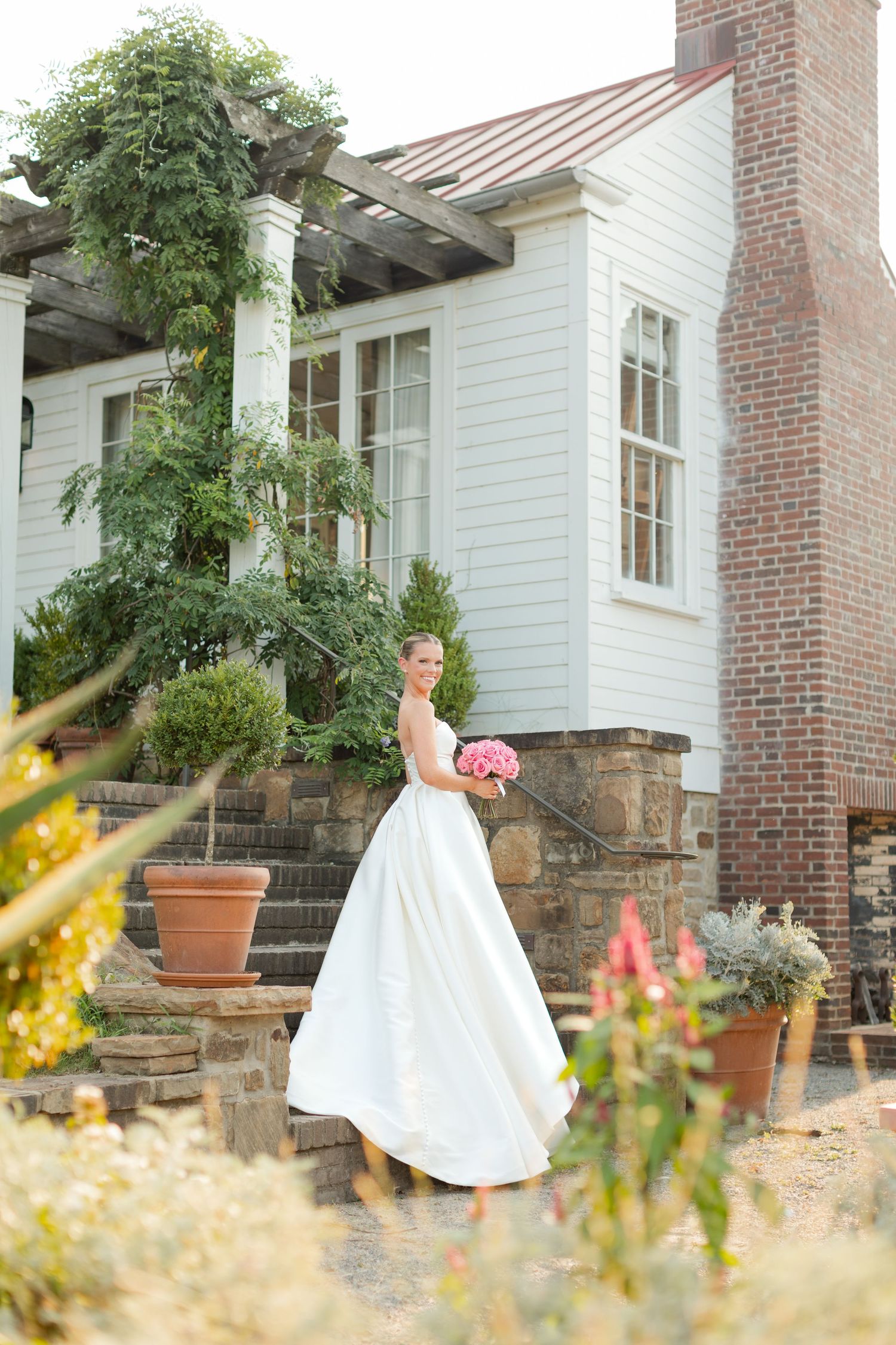 A flowing white wedding dress twirls gracefully against a rustic brick farmhouse backdrop with blooming garden flowers.
