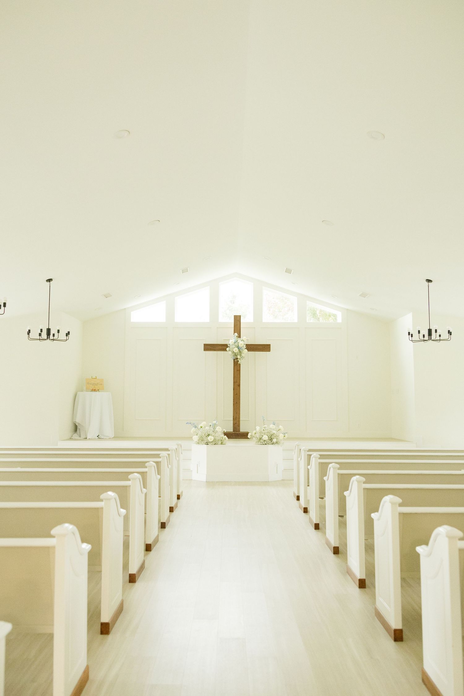 Minimalist white chapel interior with wooden cross and rows of white pews illuminated by natural light.