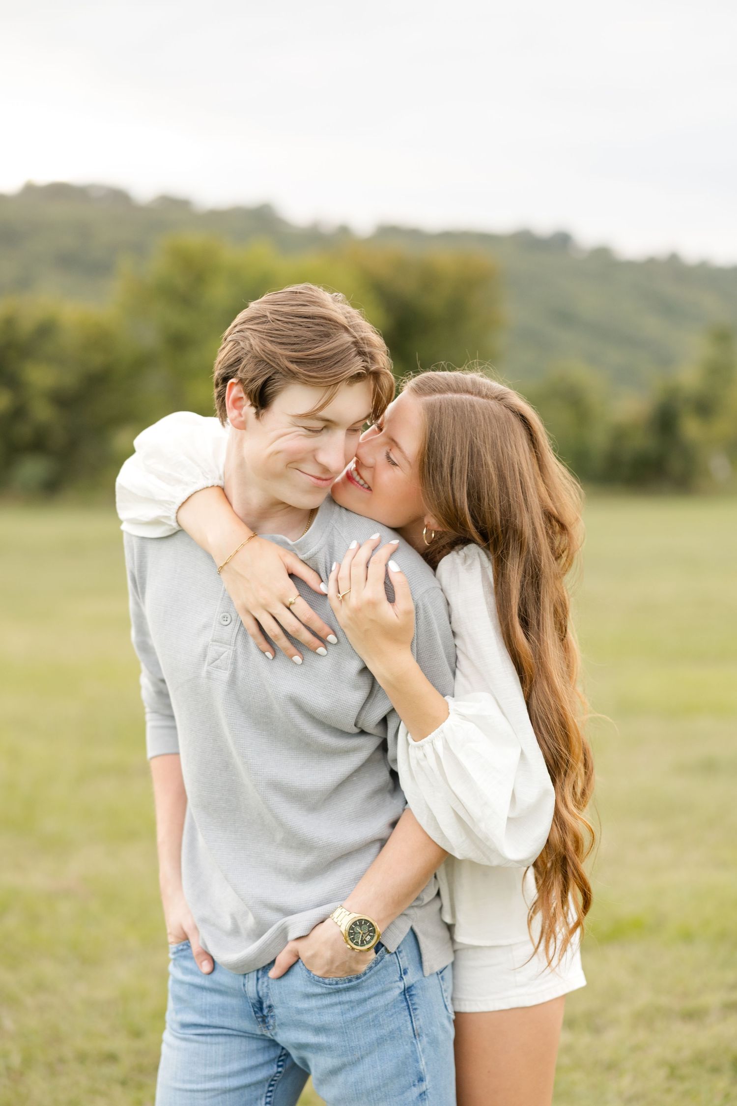 Couple sharing romantic kisses while standing in a sunny meadow with green hills in the background.