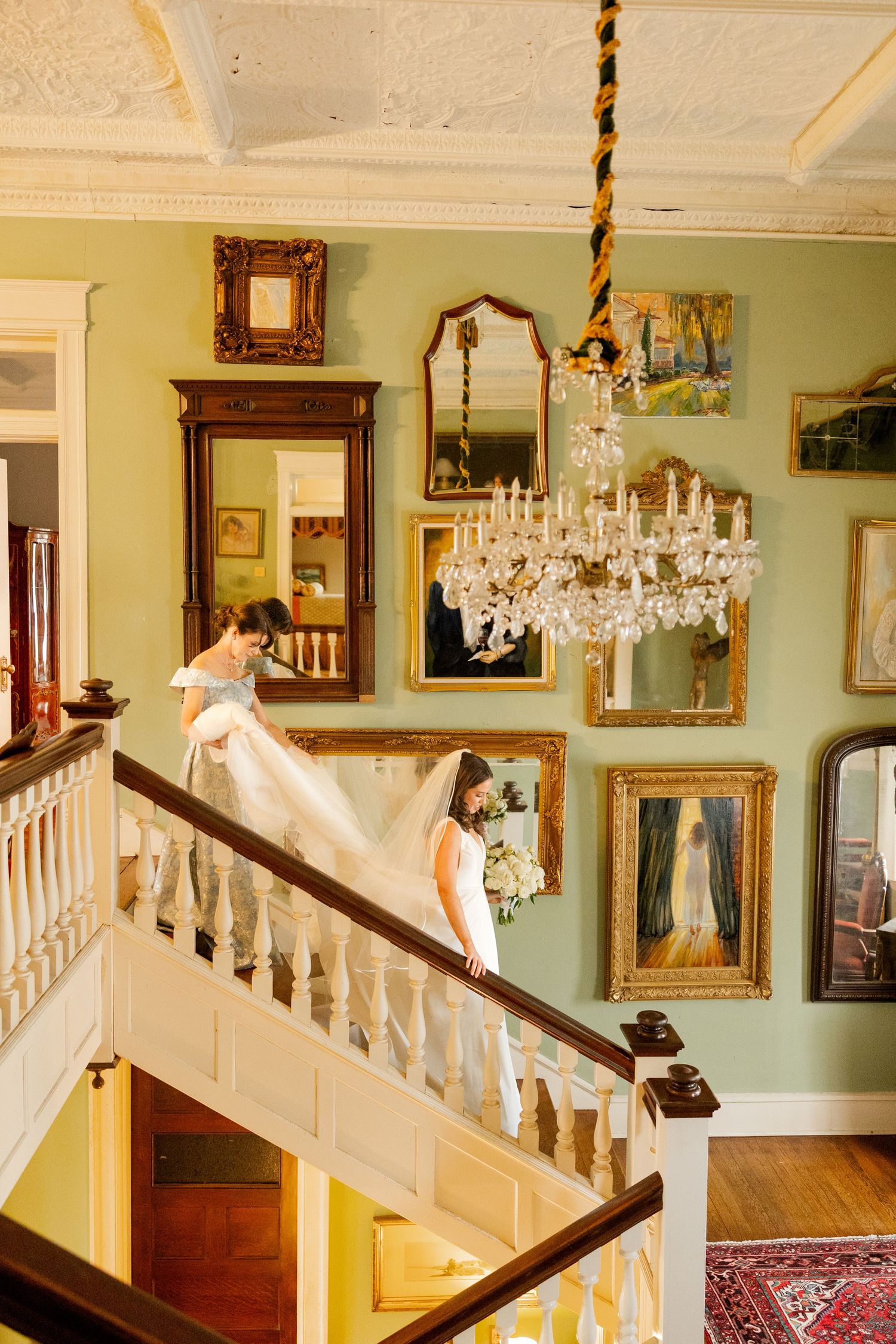 Elegant staircase in historic home with mint green walls, ornate chandelier, and vintage framed artwork.