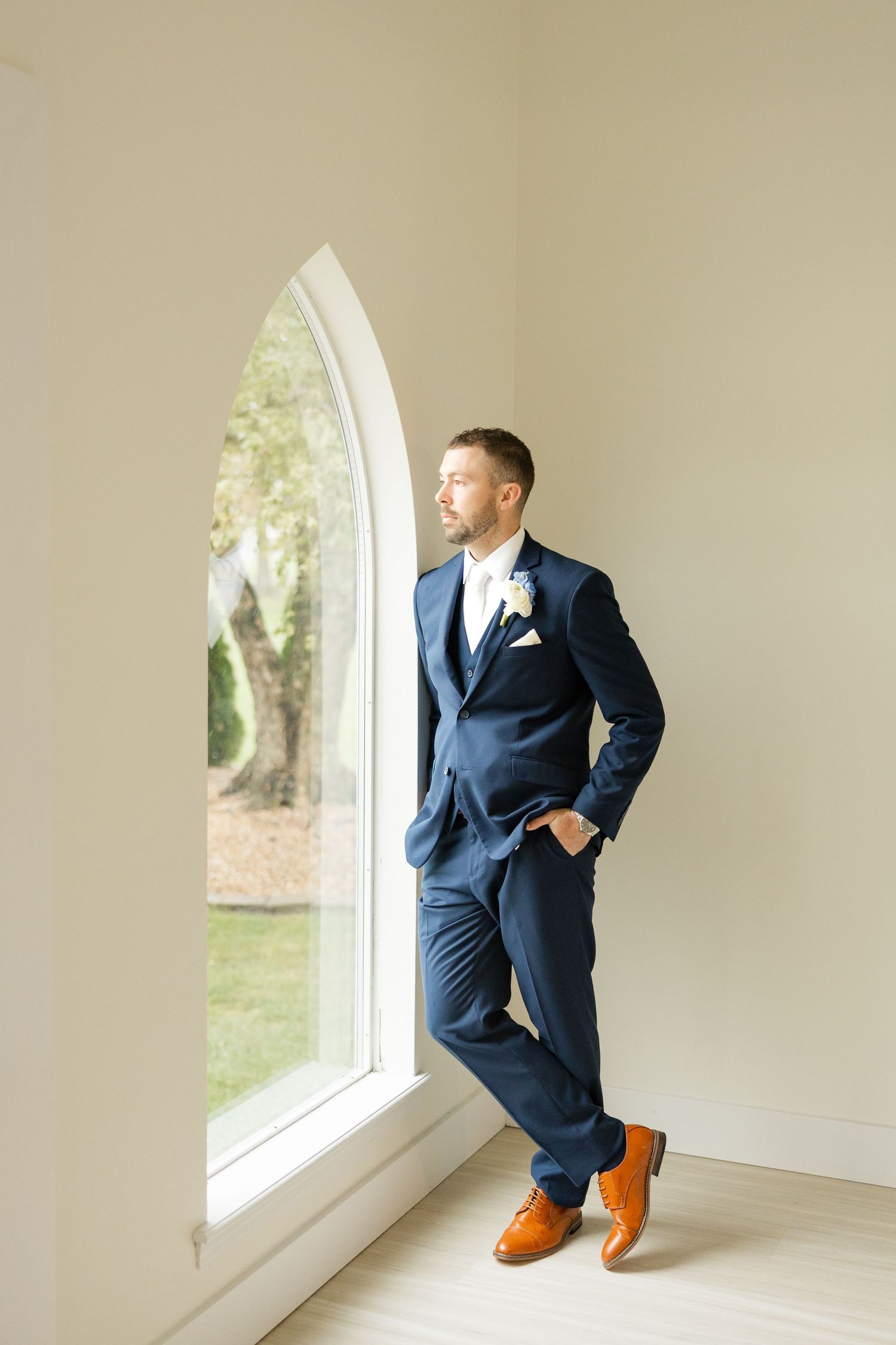 Navy blue suited figure stands by arched window in bright white room with polished hardwood floors.