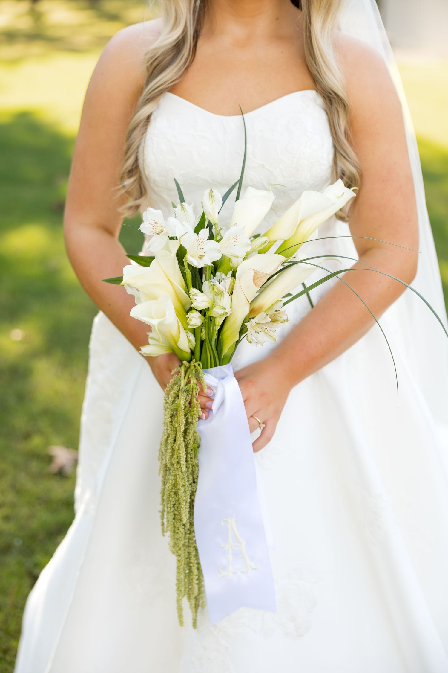 A bridal bouquet of white calla lilies and cascading amaranthus held against a flowing white wedding dress in sunlight.