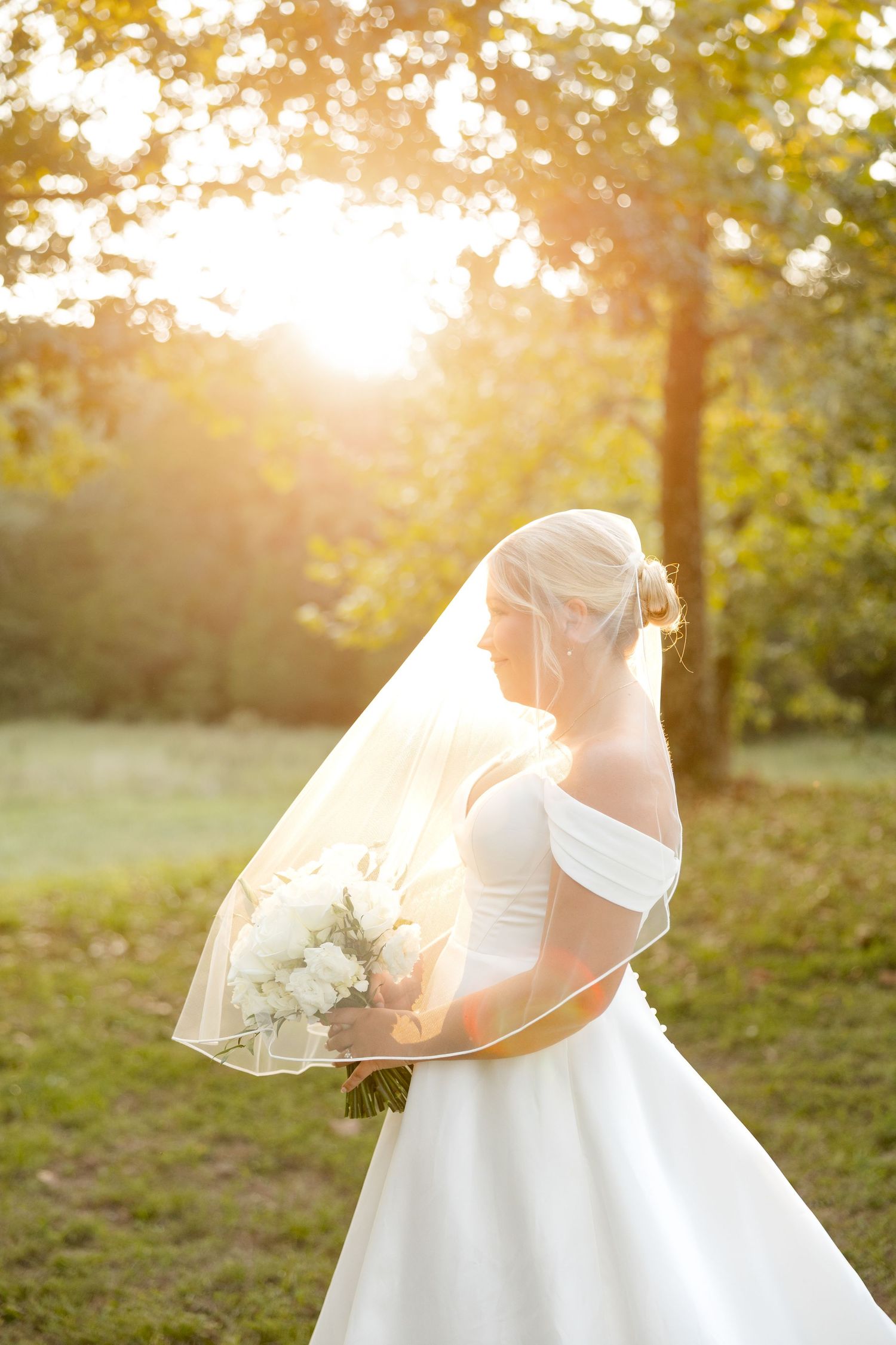 A bride in a flowing white wedding dress holds a bouquet while standing in golden sunlight during sunset.