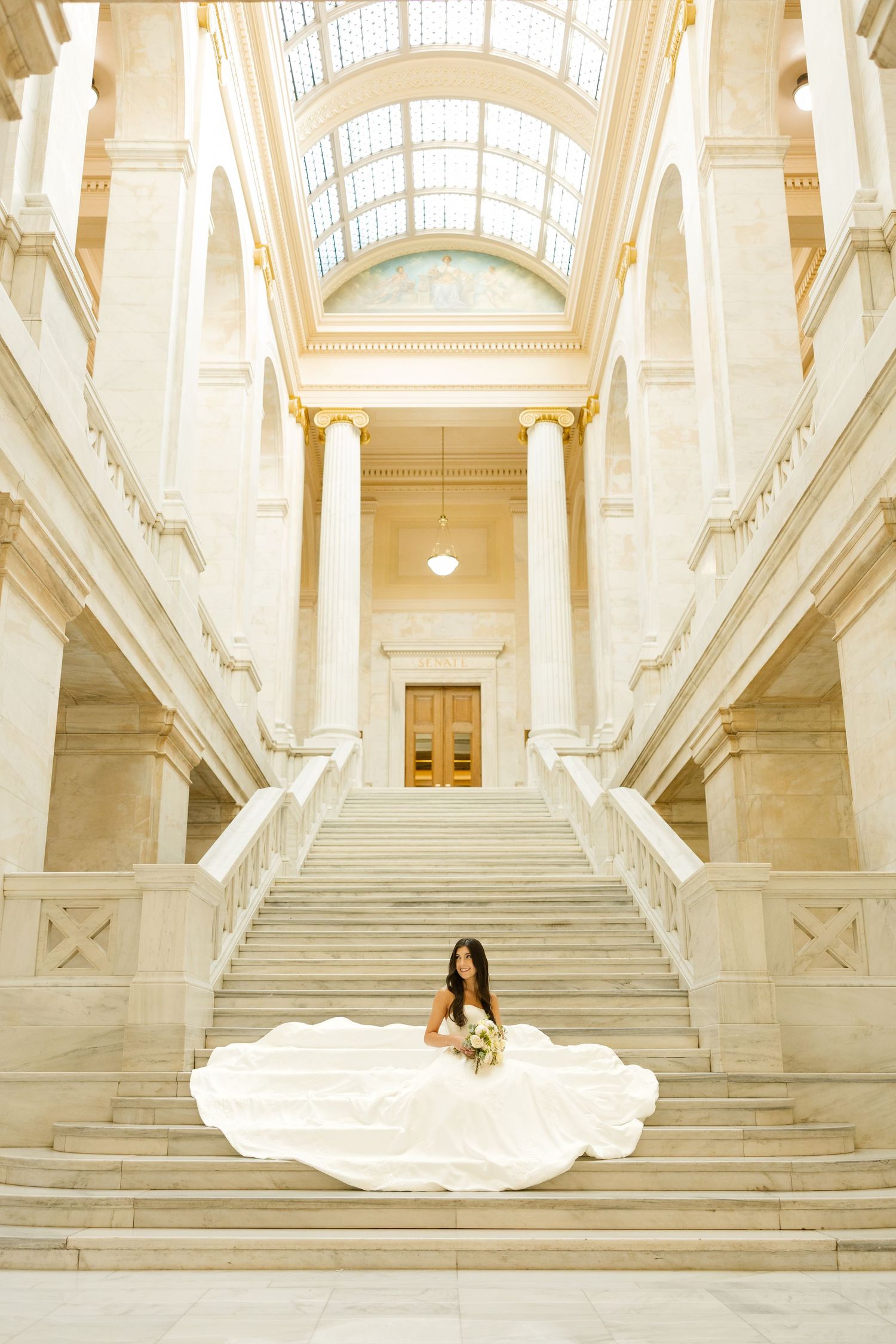 A dramatic bridal photoshoot in a grand marble hall with soaring columns and natural light streaming through windows.