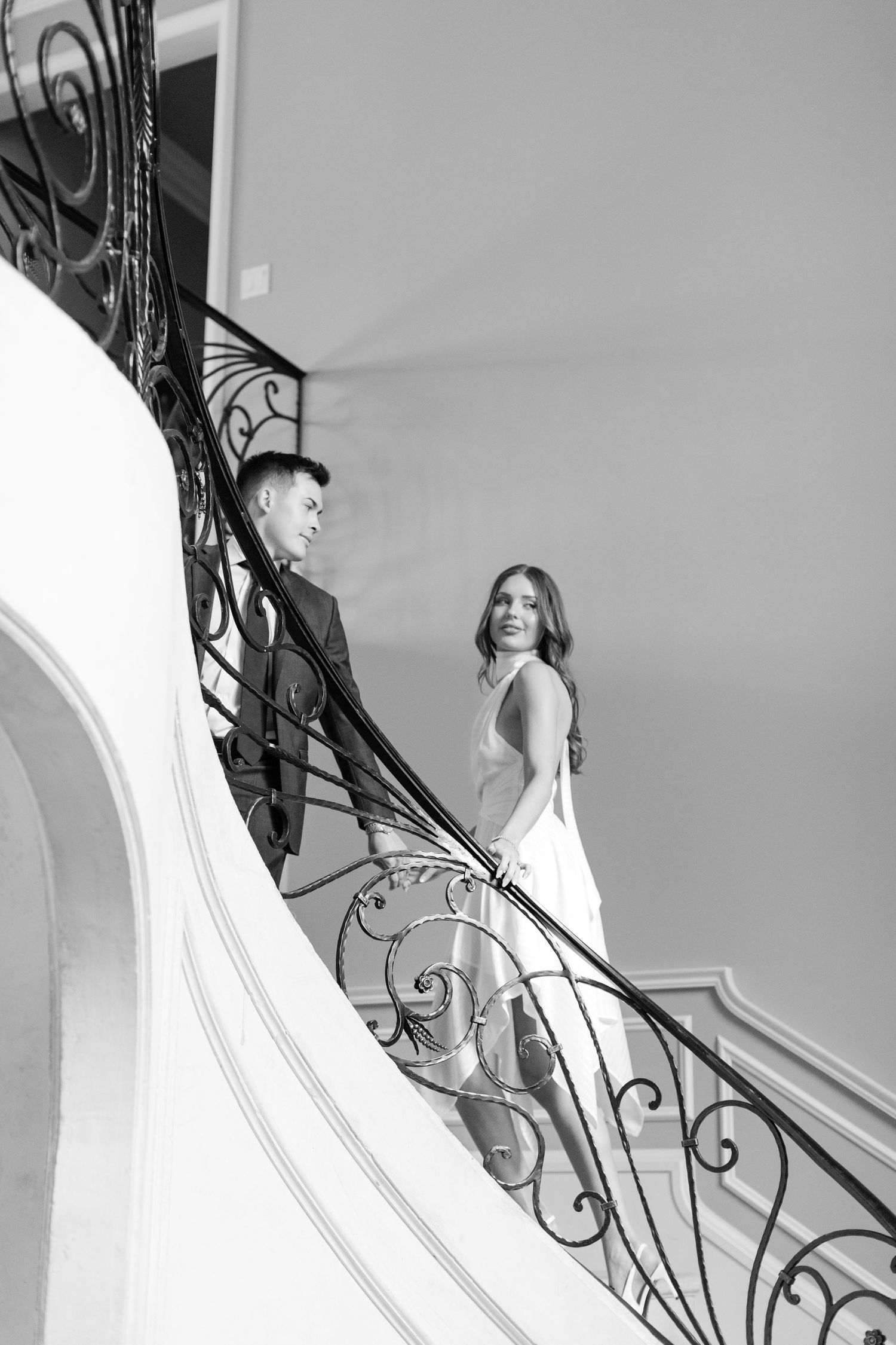 A couple shares a romantic moment on an elegant staircase with ornate wrought iron railings in black and white. Goodwin Manor.