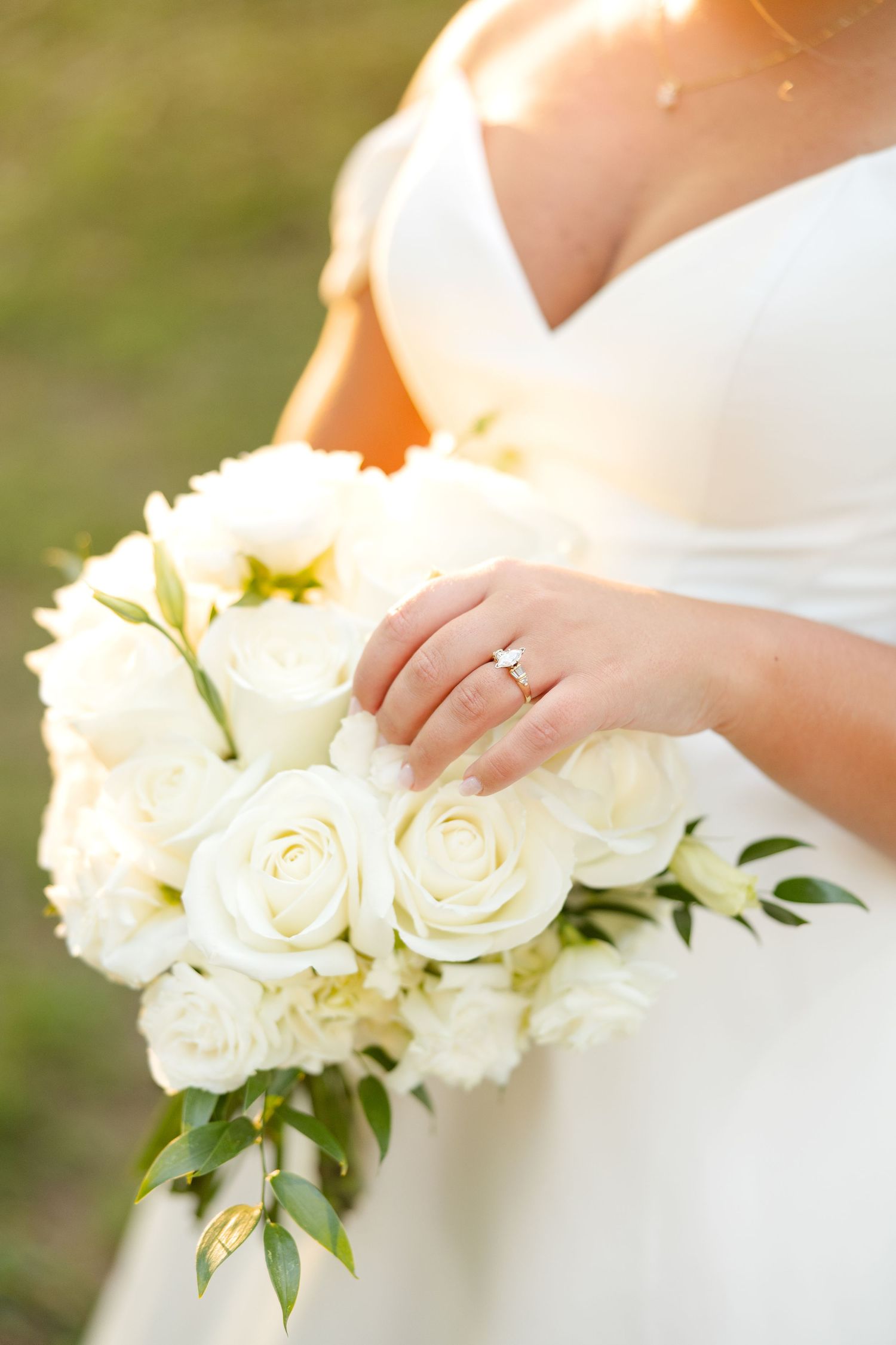 A bride in a white dress holds a classic bouquet of cream roses and greenery against a soft outdoor background.