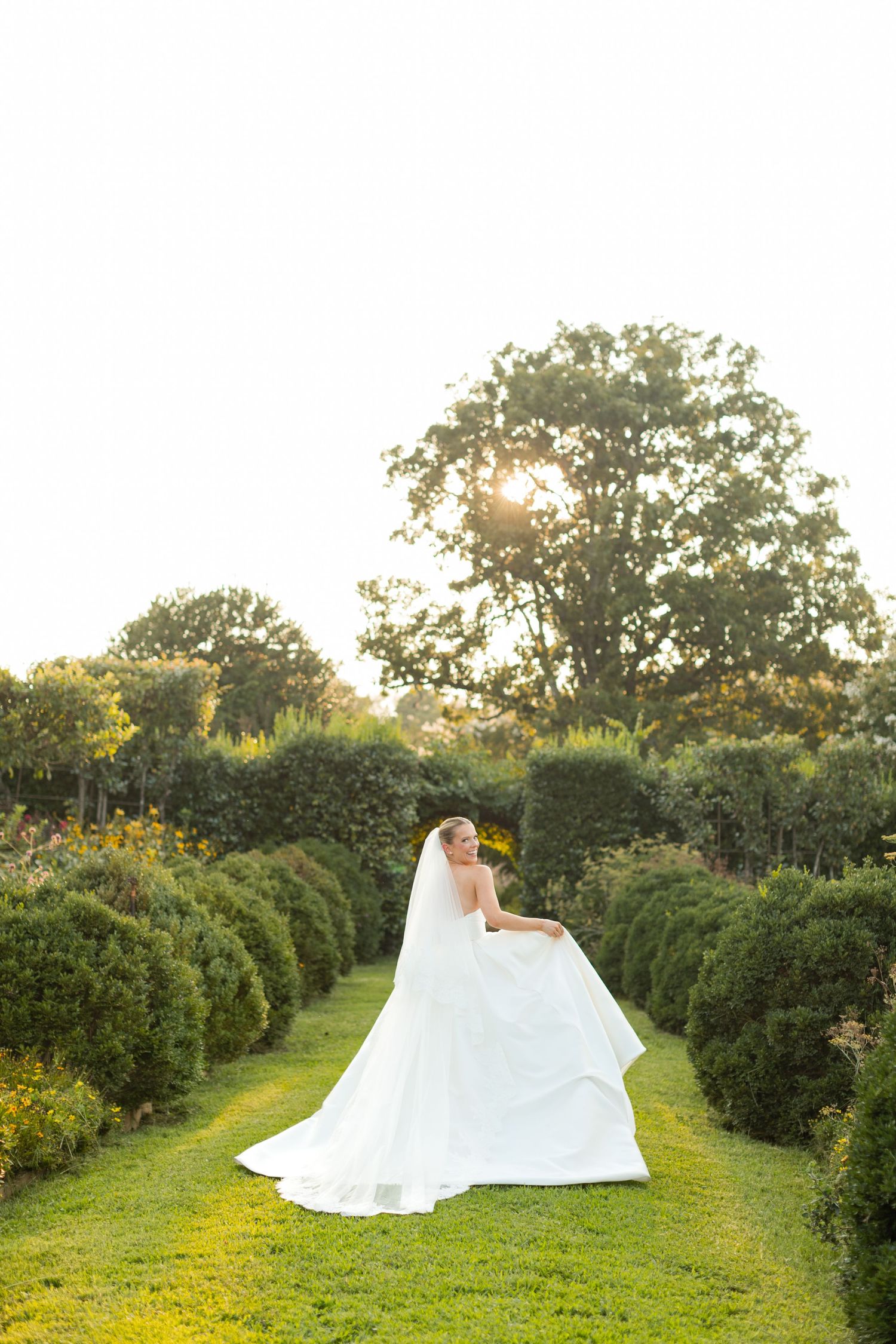 An ethereal bridal portrait shows a white wedding dress billowing across a manicured garden path at sunset.